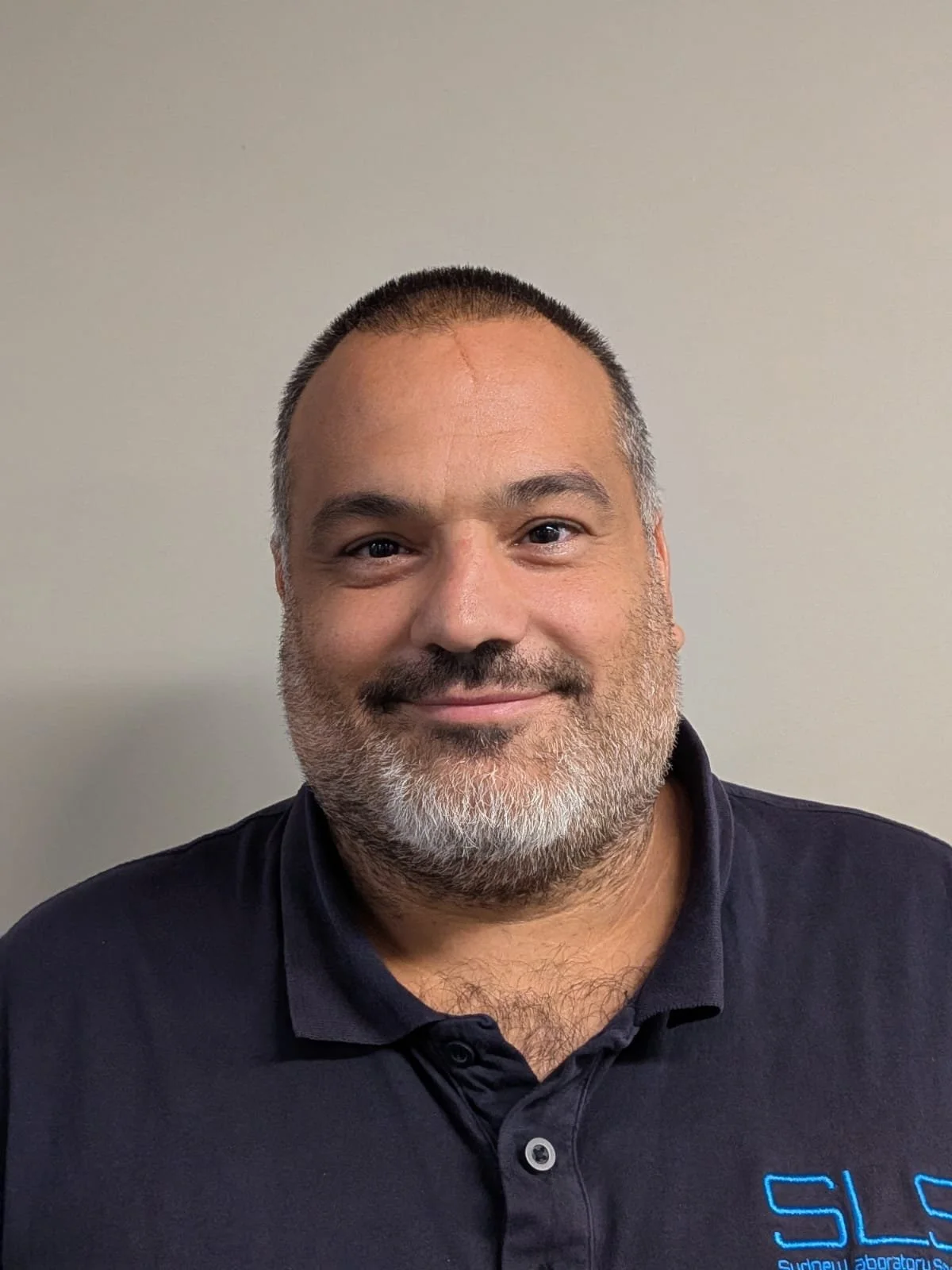A man with short dark hair, a beard, and light skin, smiling and wearing a black collared shirt in front of a plain light-colored wall.