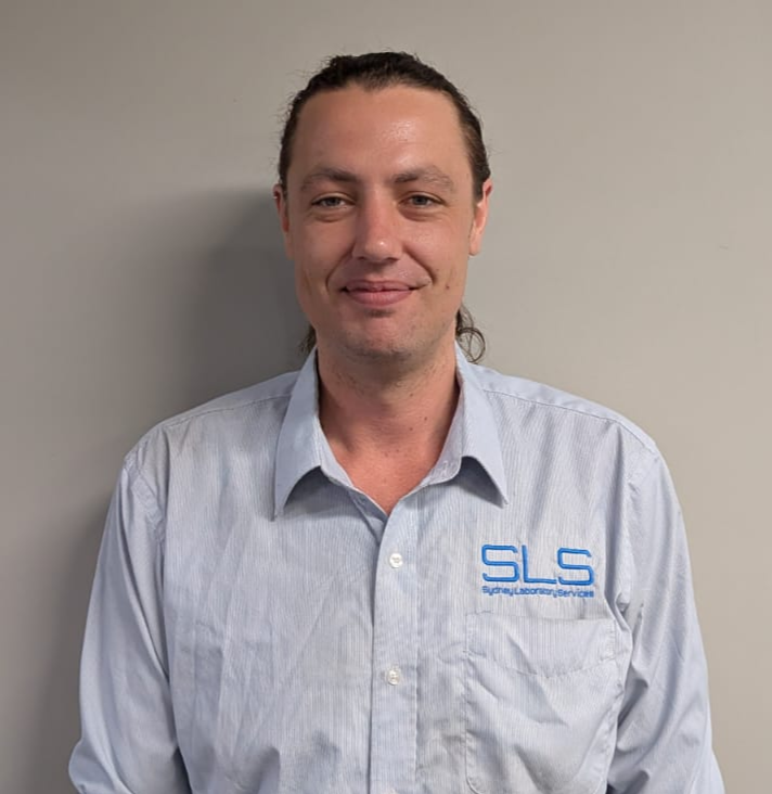 A man with long brown hair tied back, wearing a light blue dress shirt with an 'SLS' logo on the chest, standing against a plain background.