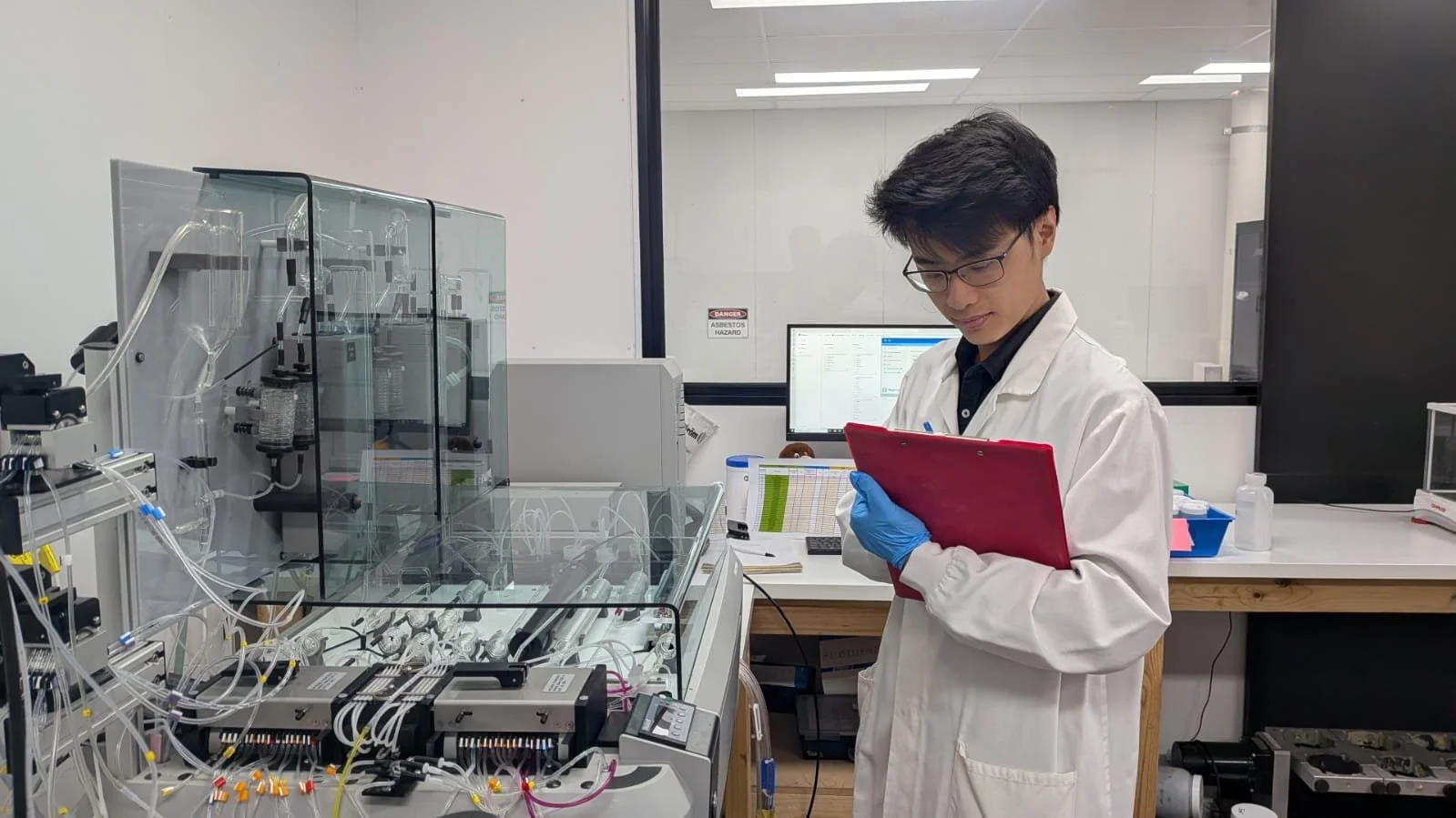 A scientist in a white lab coat and blue gloves taking notes on a red tablet in a laboratory with scientific equipment and a computer monitor in the background.