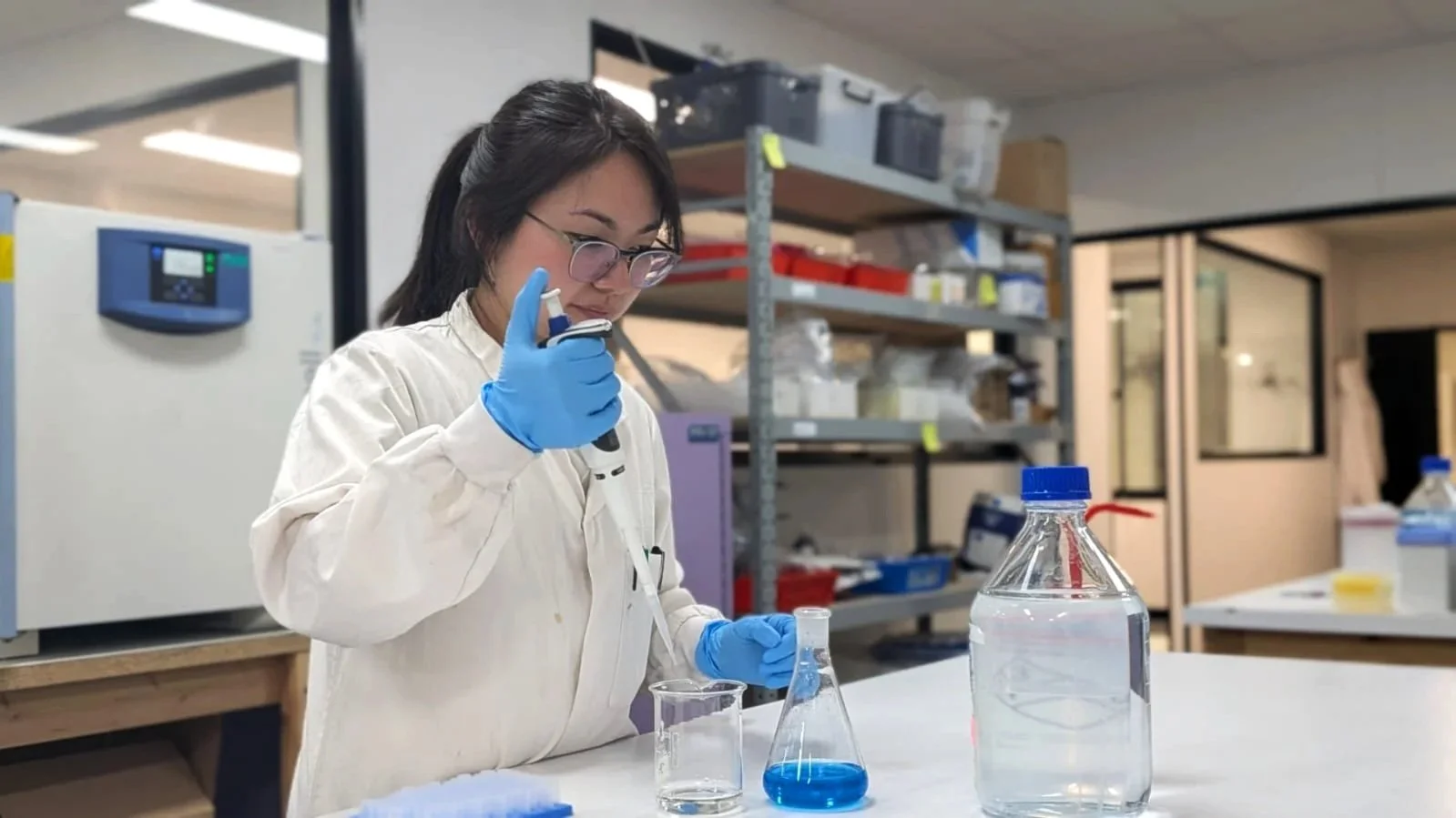 A woman wearing glasses, a white lab coat, and blue gloves is working with laboratory equipment, including a glass beaker filled with blue liquid, in a science laboratory.