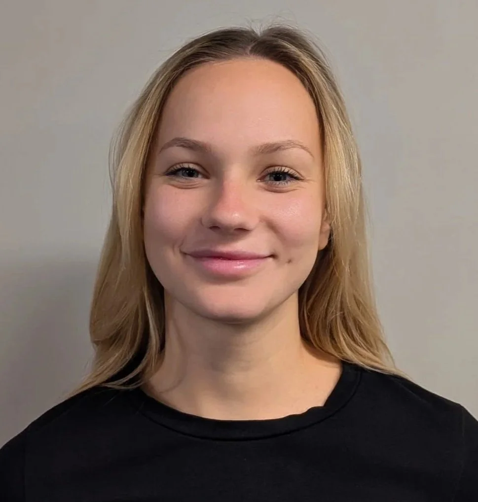 A young woman with blonde hair smiling at camera, wearing a black shirt, standing against a plain gray wall.