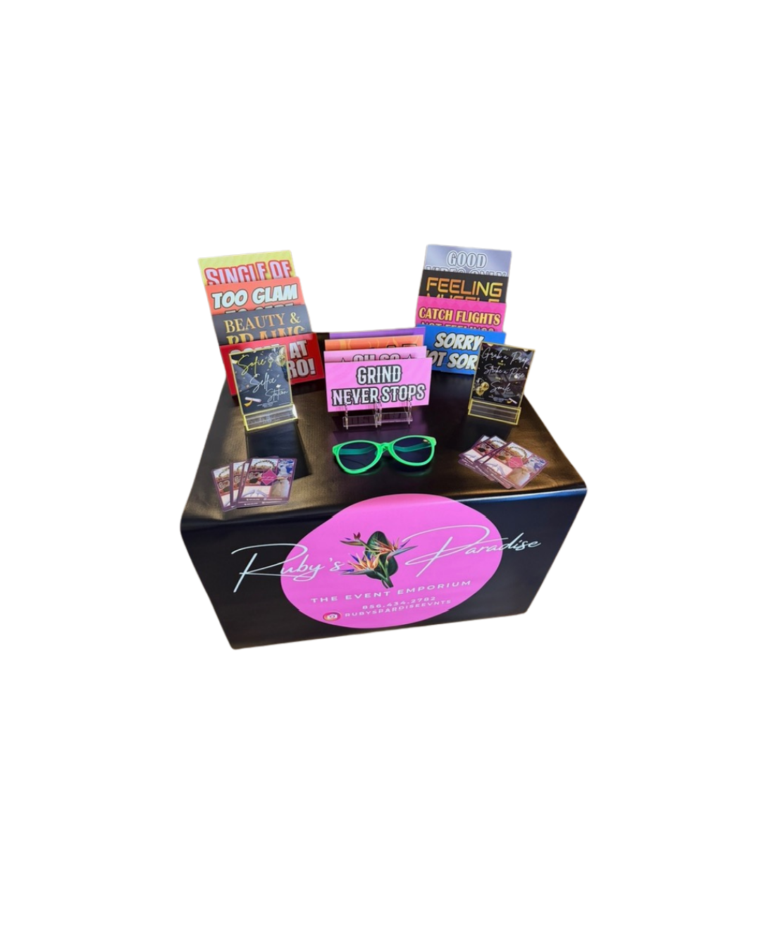 Display table with motivational signs, pink sunglasses, and promotional materials for Ruby's Paradise event at The Event Emporium.