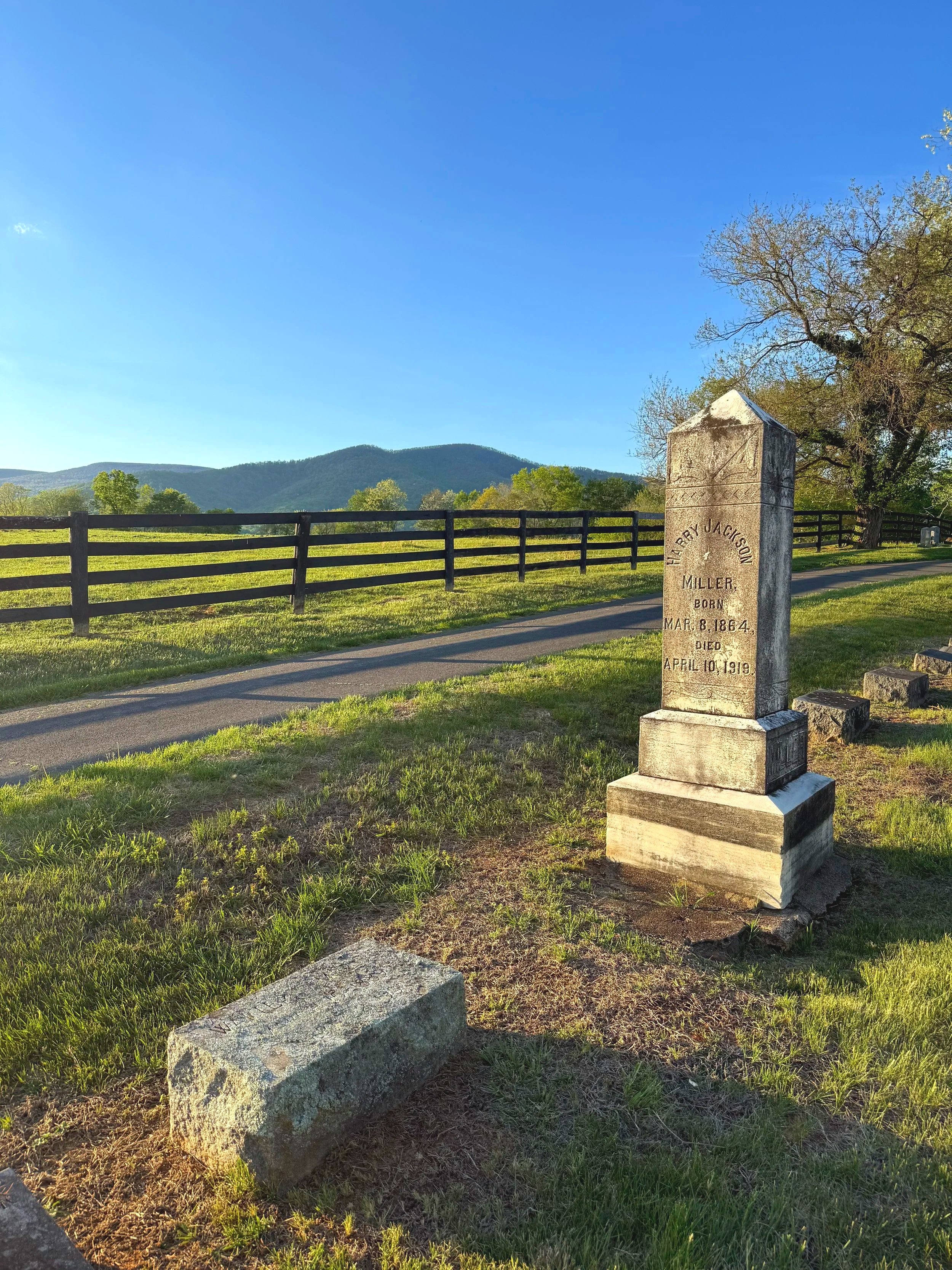 A stone gravestone with inscriptions in front of a park bench, on a grassy area with a fence, trees, and mountains in the background during late afternoon.