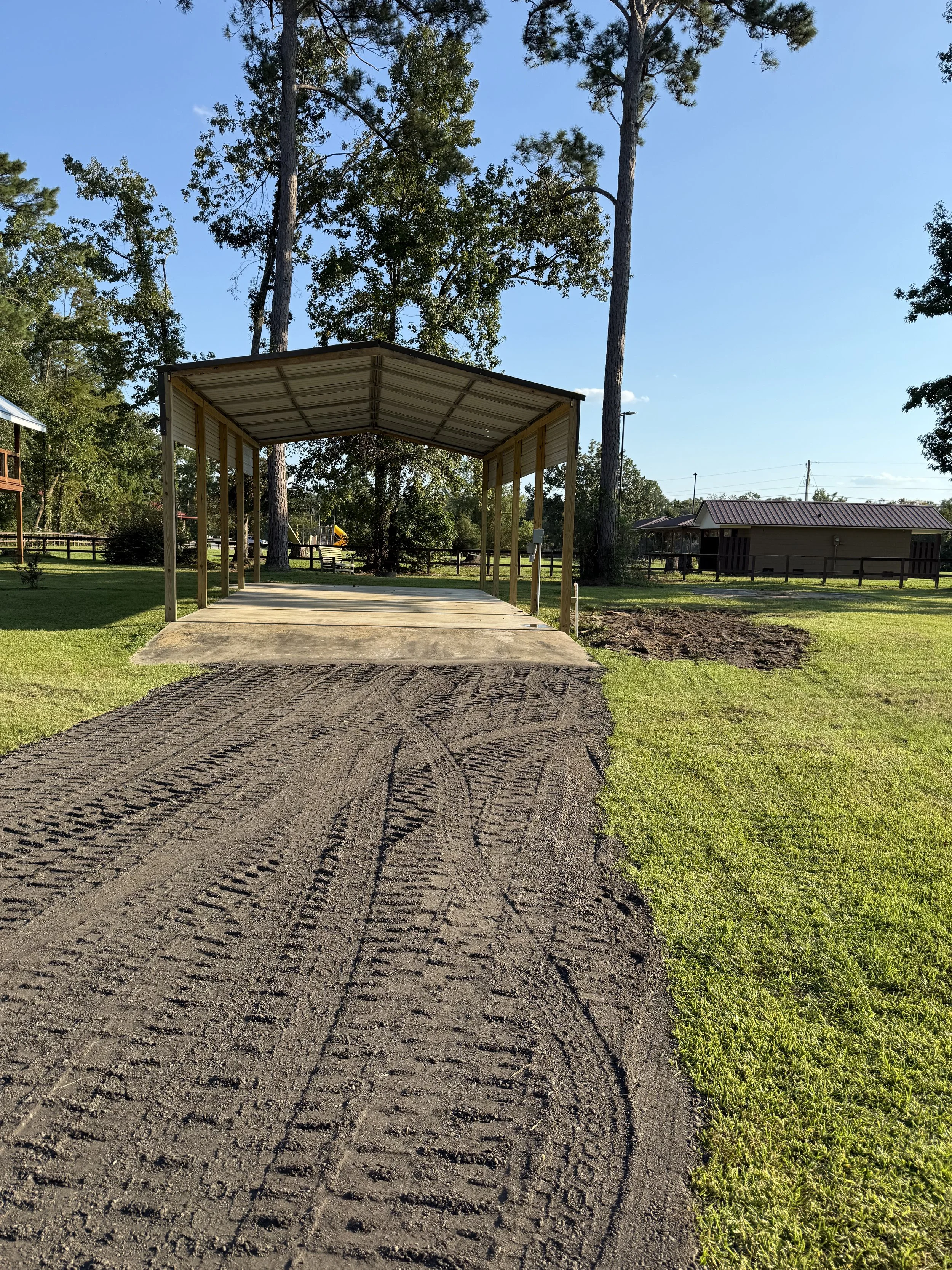 A dirt driveway leading to a covered parking area with a metal roof, surrounded by green grass and tall trees in the background.