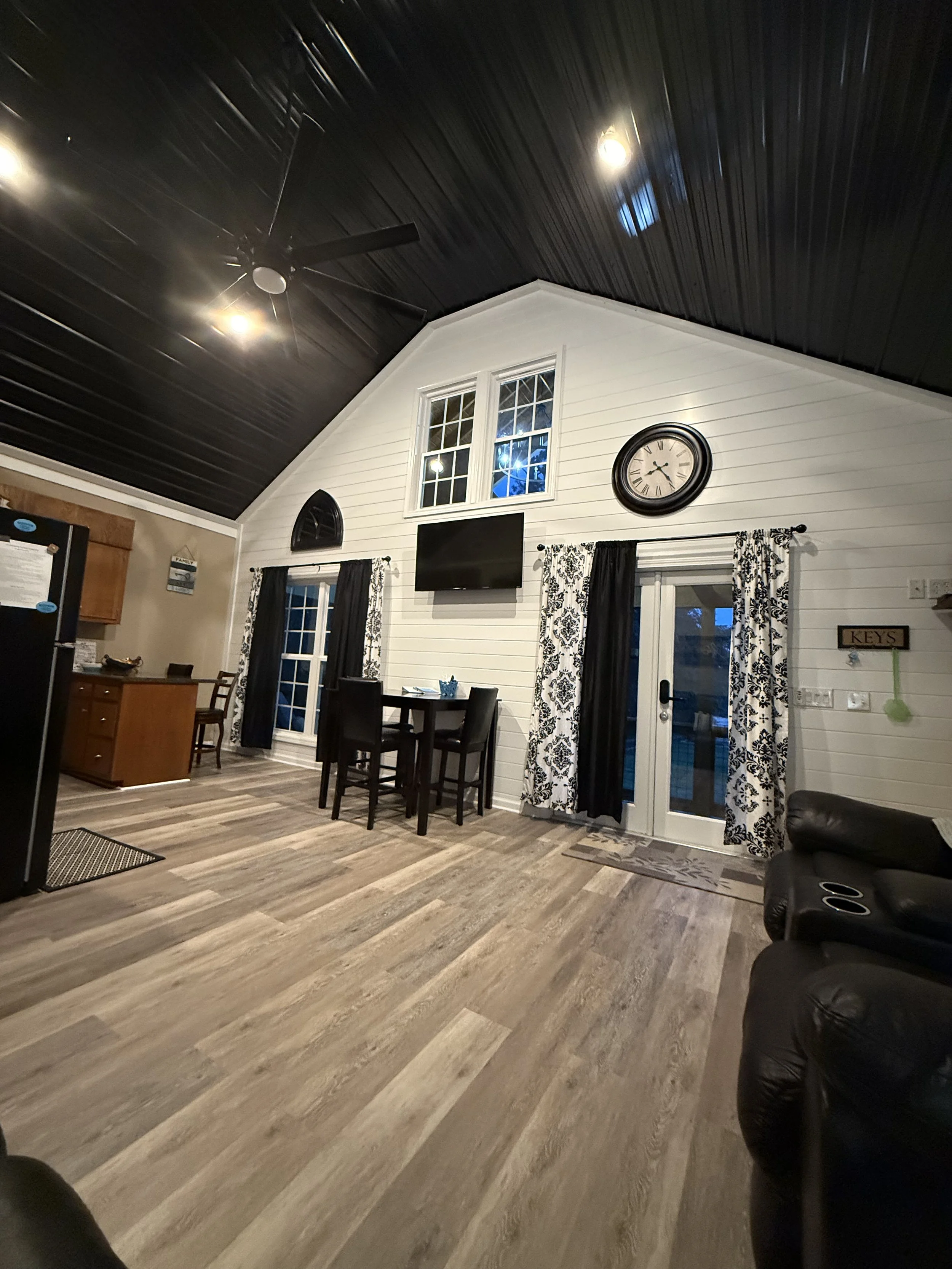 Living room with high vaulted black ceiling, wood flooring, white shiplap walls, black and white patterned curtains, a small dining table with four chairs, a wall-mounted TV, a large wall clock, sliding glass door, and a partial view of kitchen area.