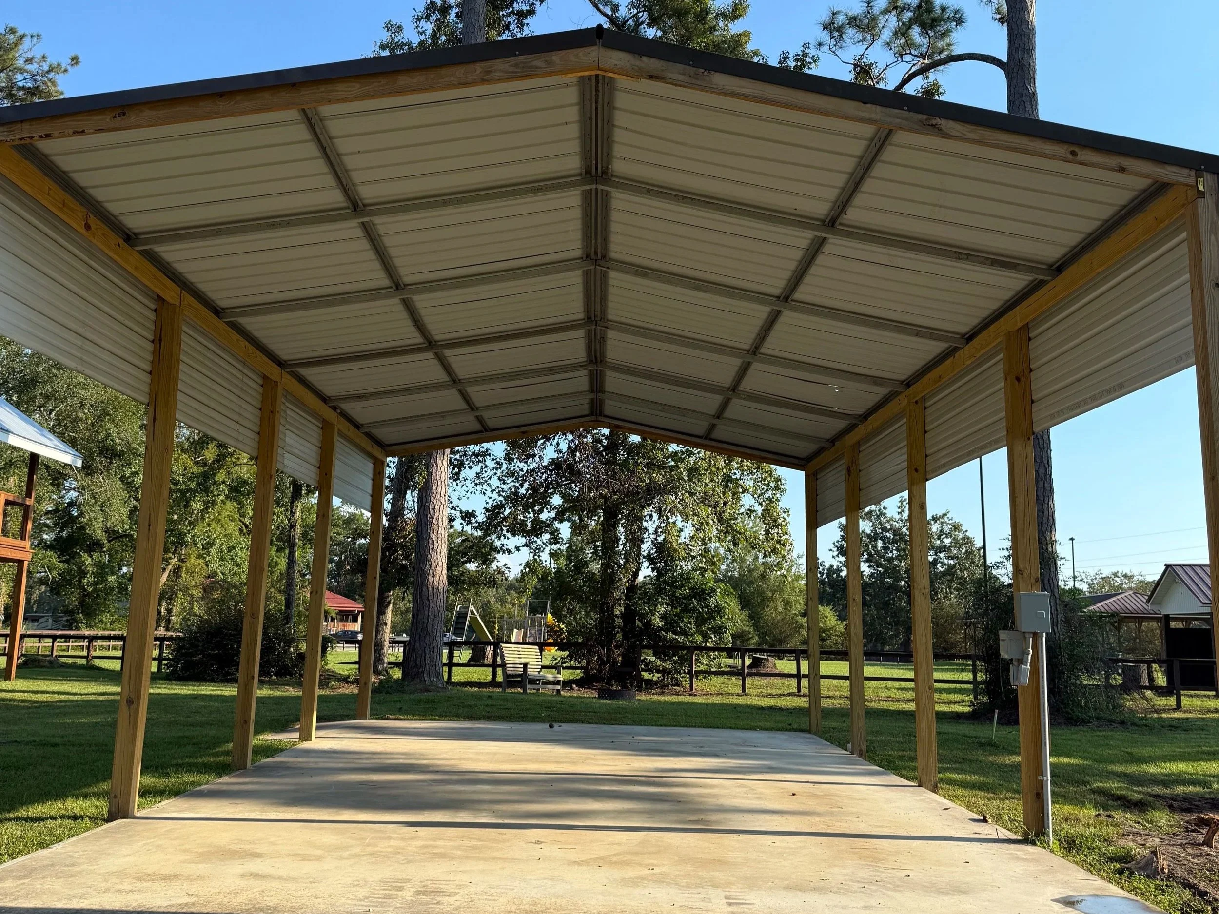 A partially built outdoor shelter with a metal roof and wooden frame on a concrete slab, surrounded by green grass and trees.