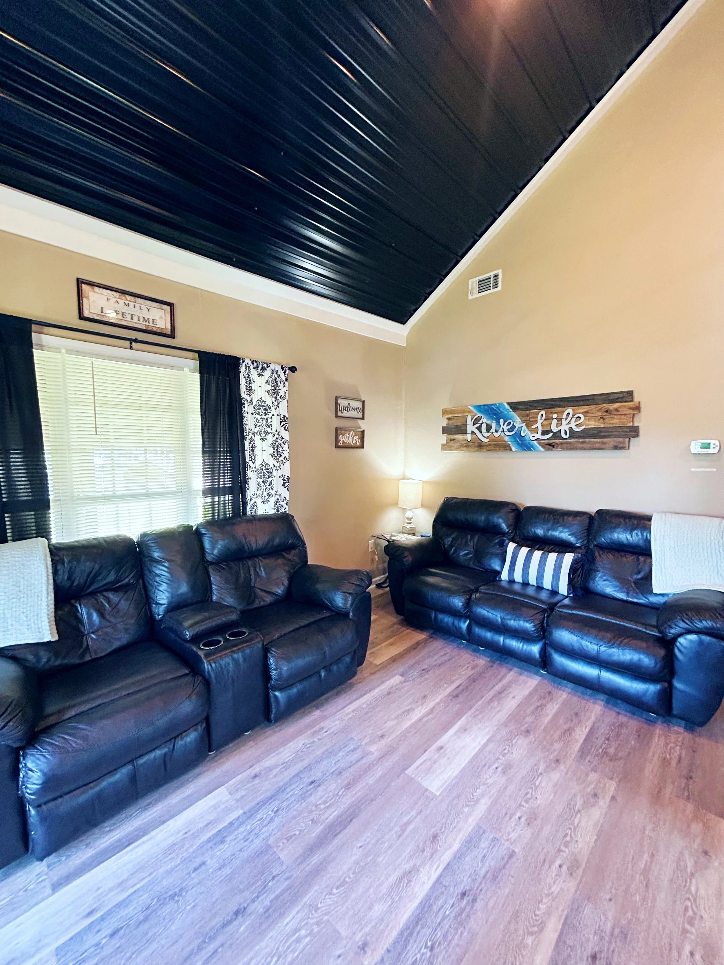 Living room with two black leather sofas, a window with black and white patterned curtains, a beige wall with a rustic 'River Life' sign, a small lamp, and decorative wall signs that say 'welcome' and 'gather'.