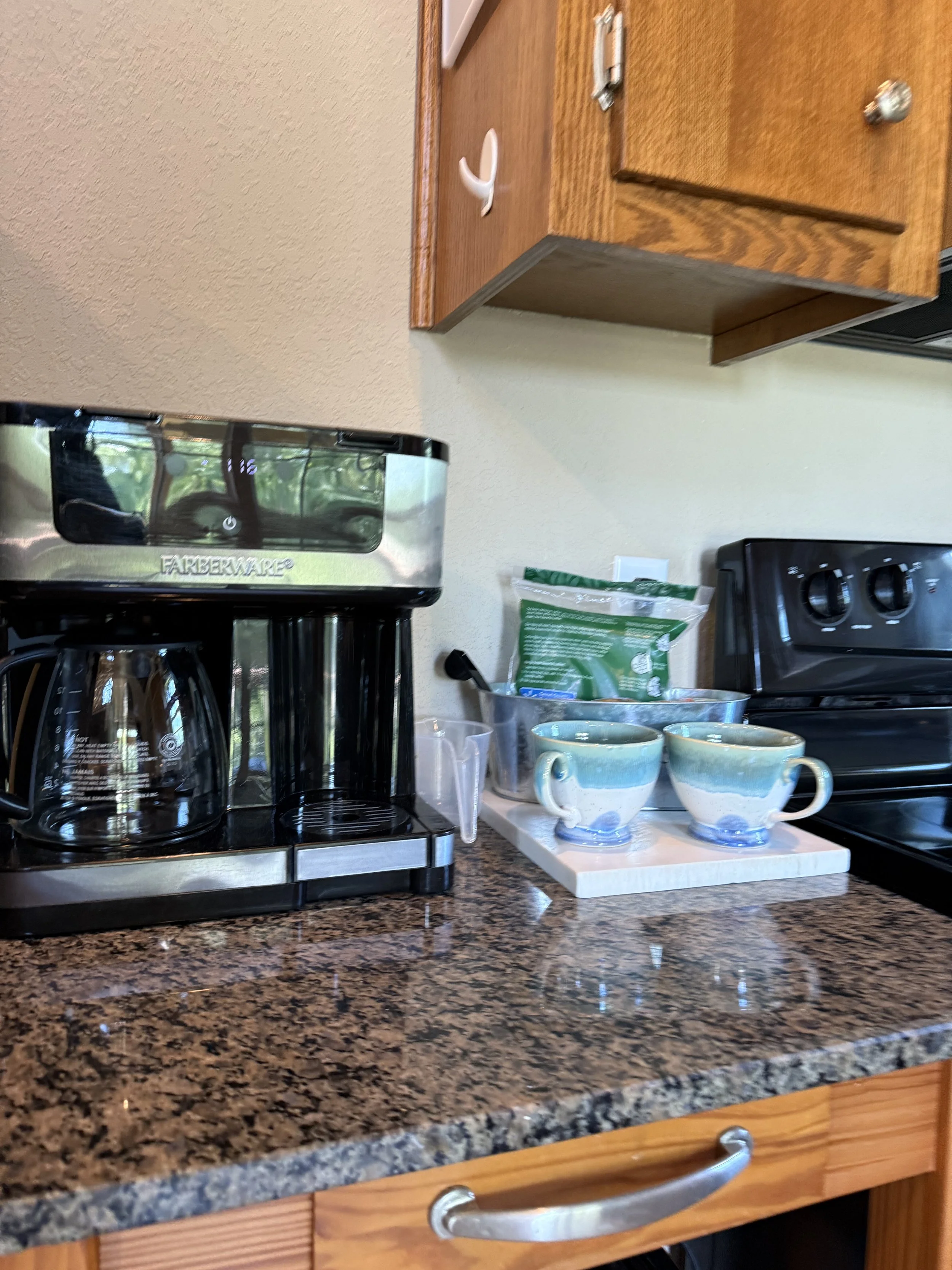 Kitchen countertop with a coffee maker, two ceramic coffee cups on a white tray, and various kitchen items. Wooden cabinets above.