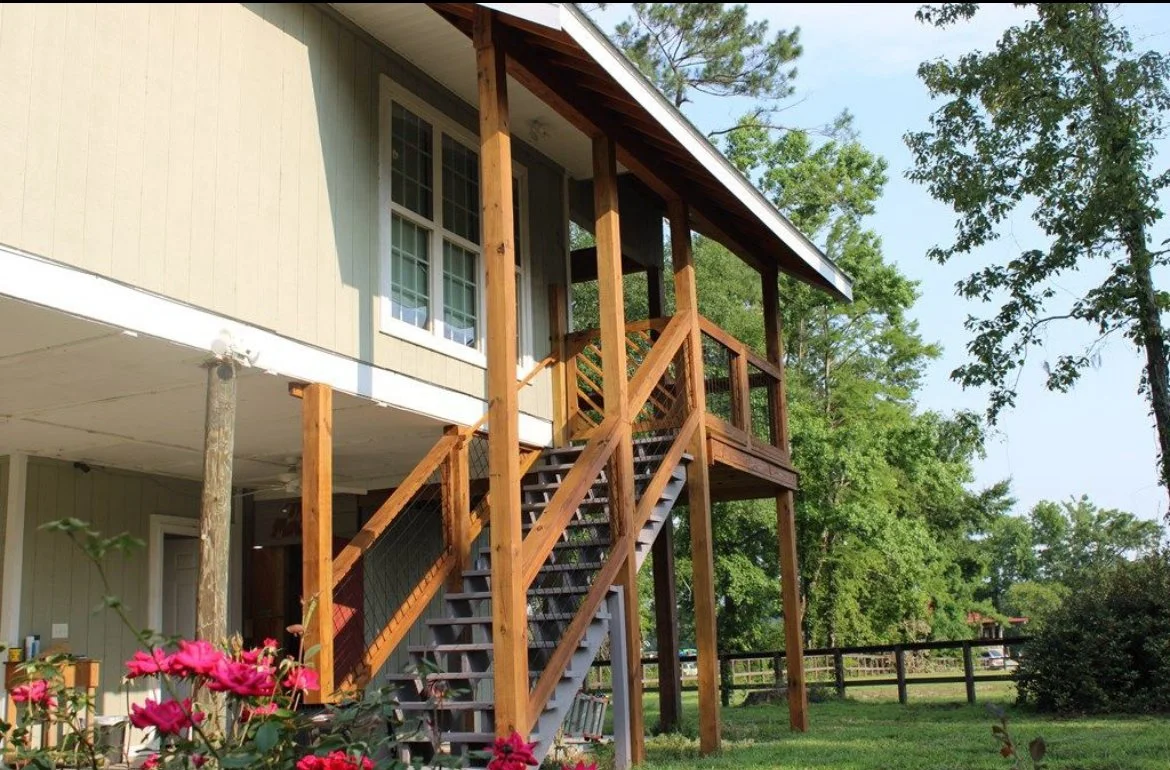 Rear view of a two-story house with a wooden staircase and balcony, surrounded by green trees and pink flowers in the foreground.