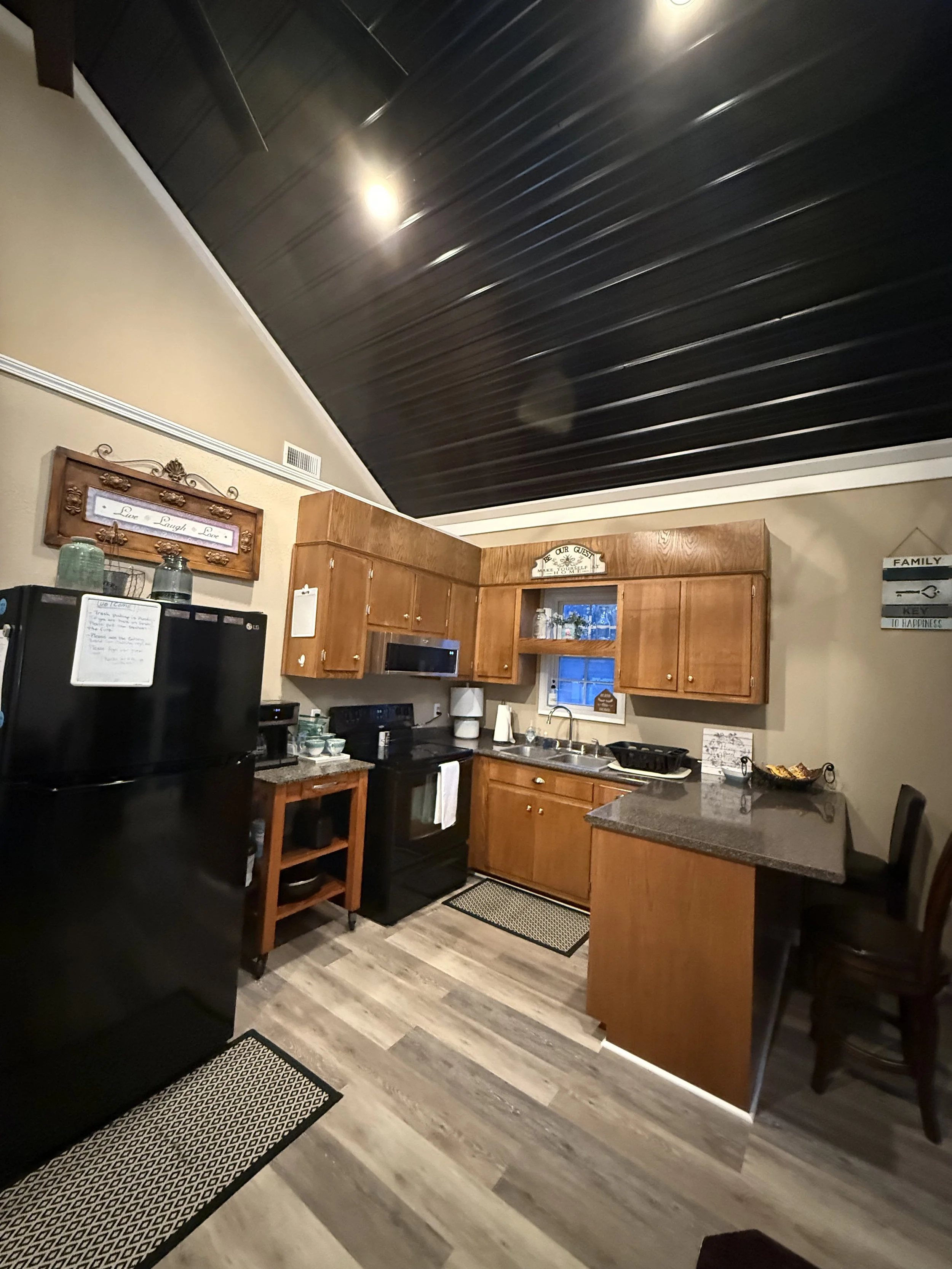 Kitchen with black fridge, black stove, wooden cabinets, small window, and gray flooring, under a dark sloped ceiling.