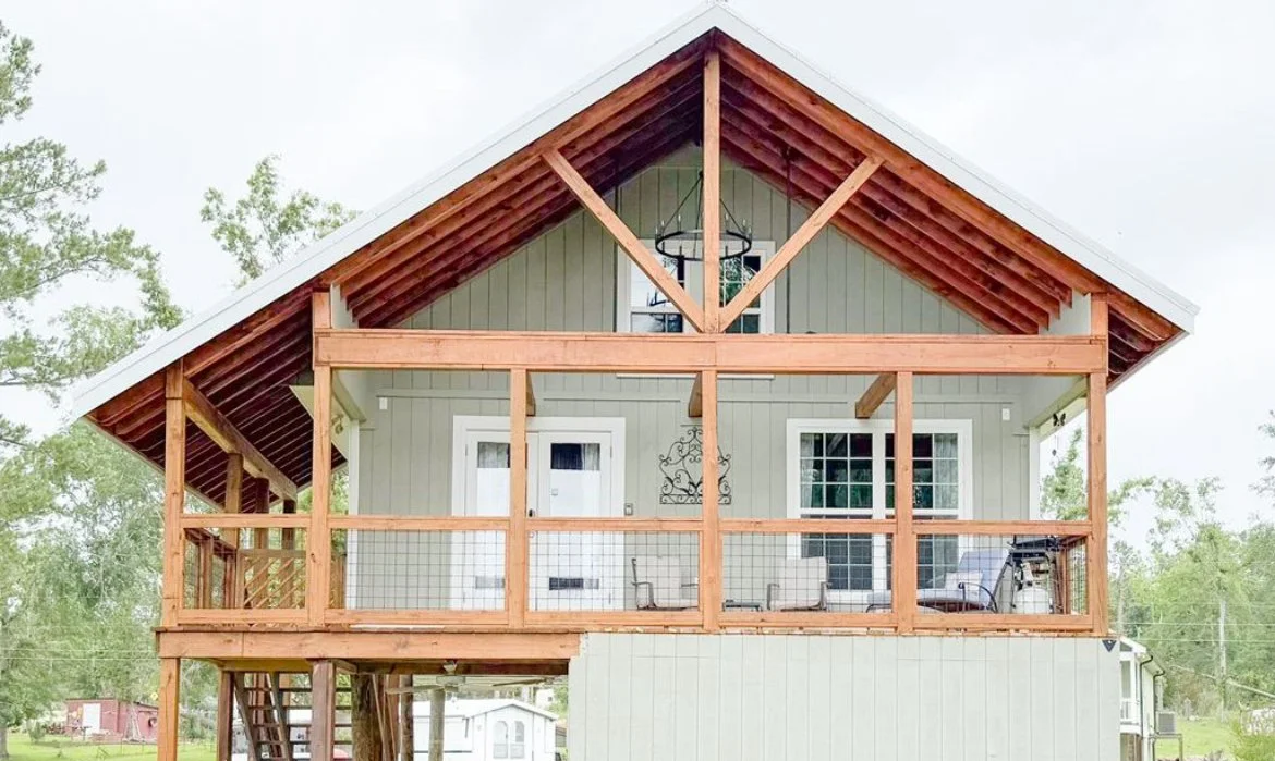 The image shows the front of a house with a wooden porch under construction. The house is painted light green with white trim, and the porch railing is installed but not yet finished. The roof has exposed wooden beams, and the background includes trees and a cloudy sky.