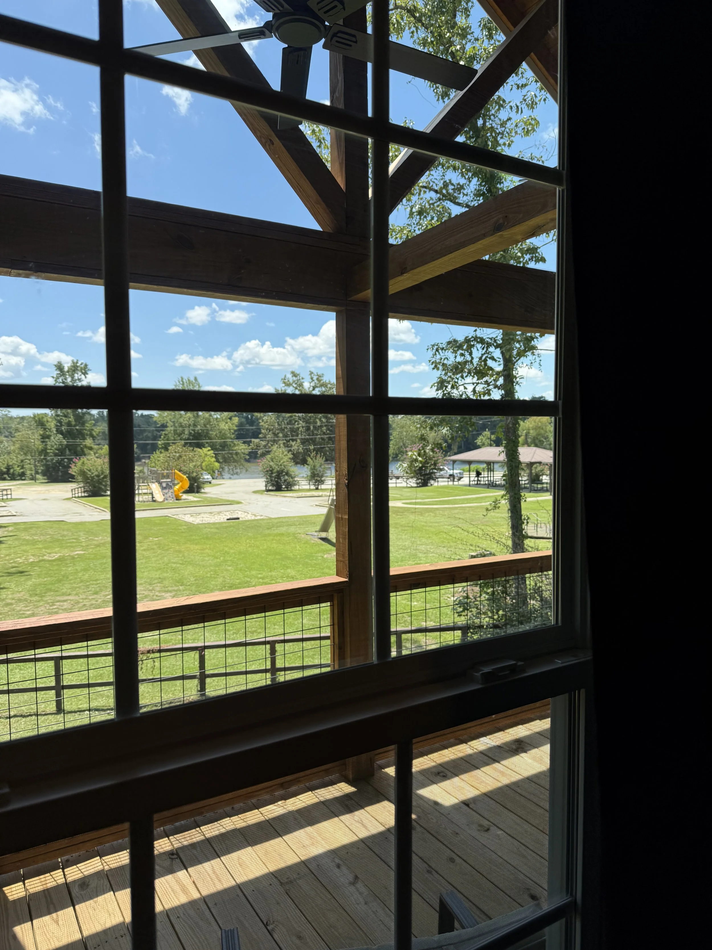 View through a window of a green park with trees, a slide, a gazebo, and a pond with a lake in the background, sunny sky with scattered clouds.