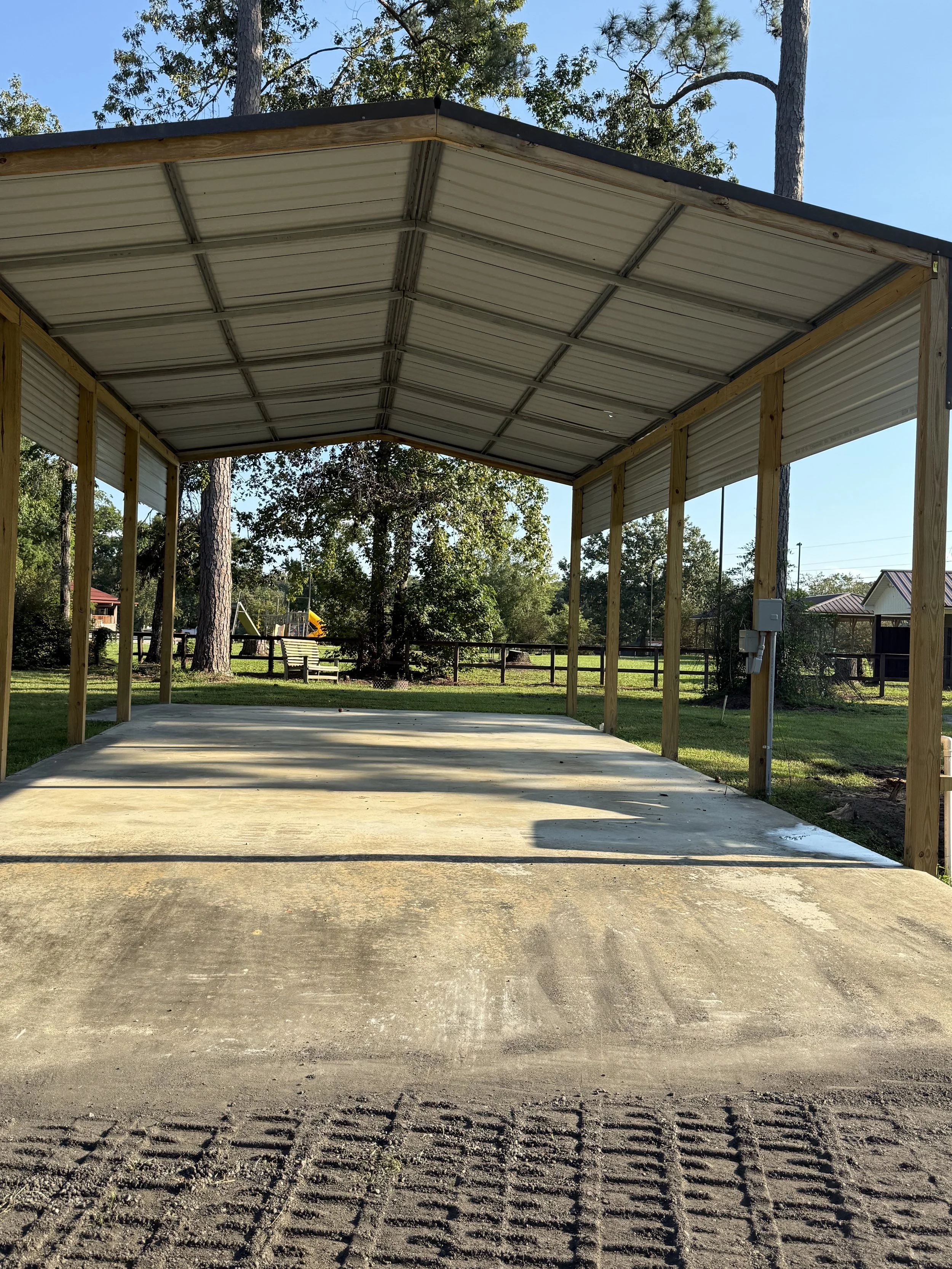 A newly constructed carport with a metal roof, wooden supporting beams, and a concrete floor located in a backyard with green grass and trees.