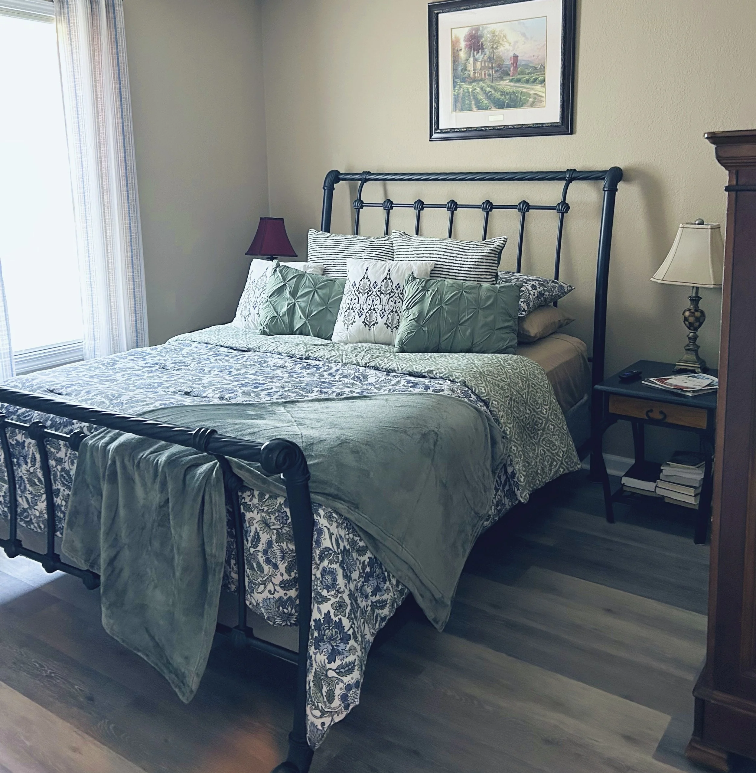 A neatly made bed with black metal frame, multiple pillows, and a patterned quilt in a bedroom with beige walls and a window with curtains. There are two nightstands with lamps on either side of the bed, and a painting hanging above the headboard.