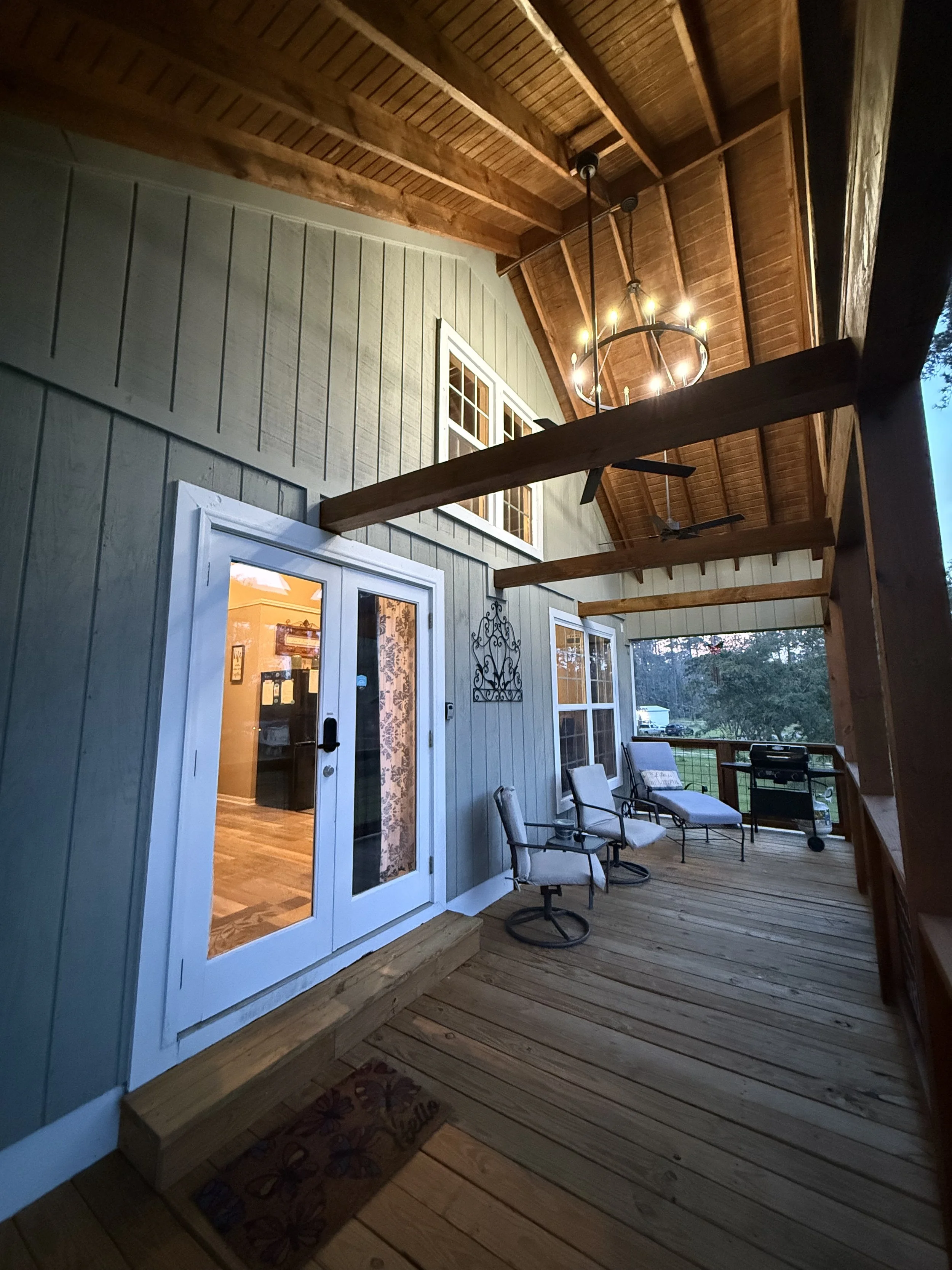 Covered outdoor porch of a house with a wooden ceiling, chandelier, and seating area including chairs and a grill, next to a sliding glass door.