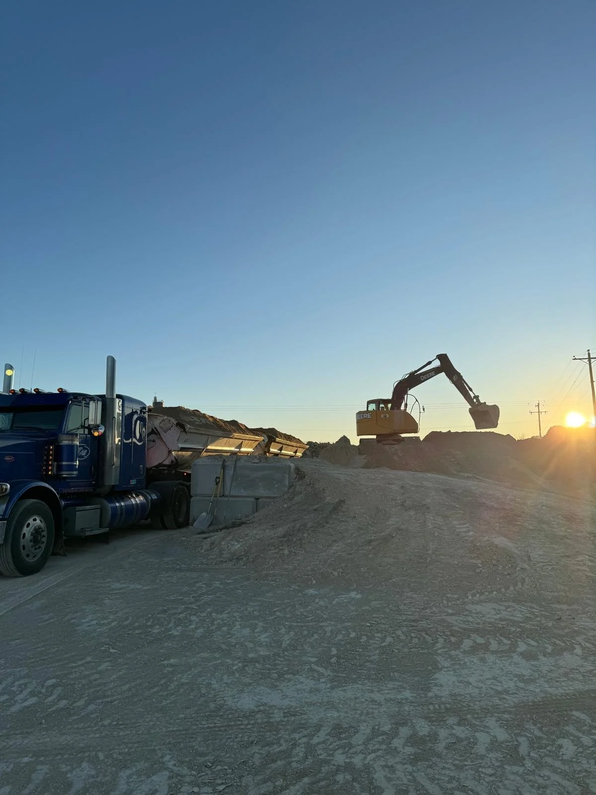 Construction site with a yellow excavator loading a blue semi-truck with lime during sunset.