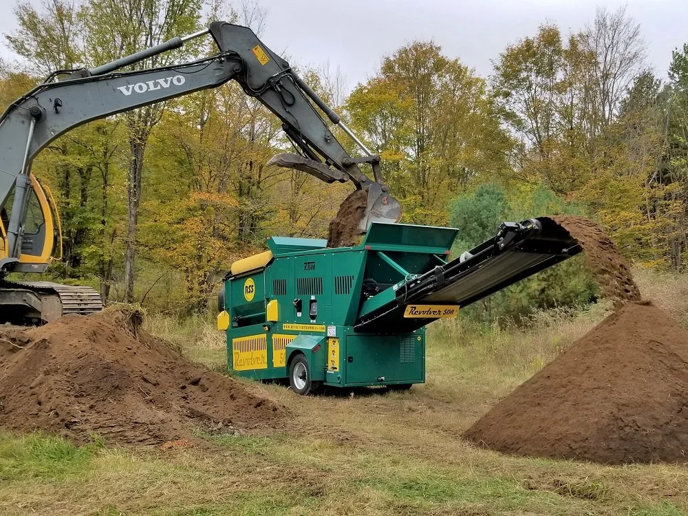 A large excavator loading soil into a green industrial soil screening machine outdoors in a forested area during fall.