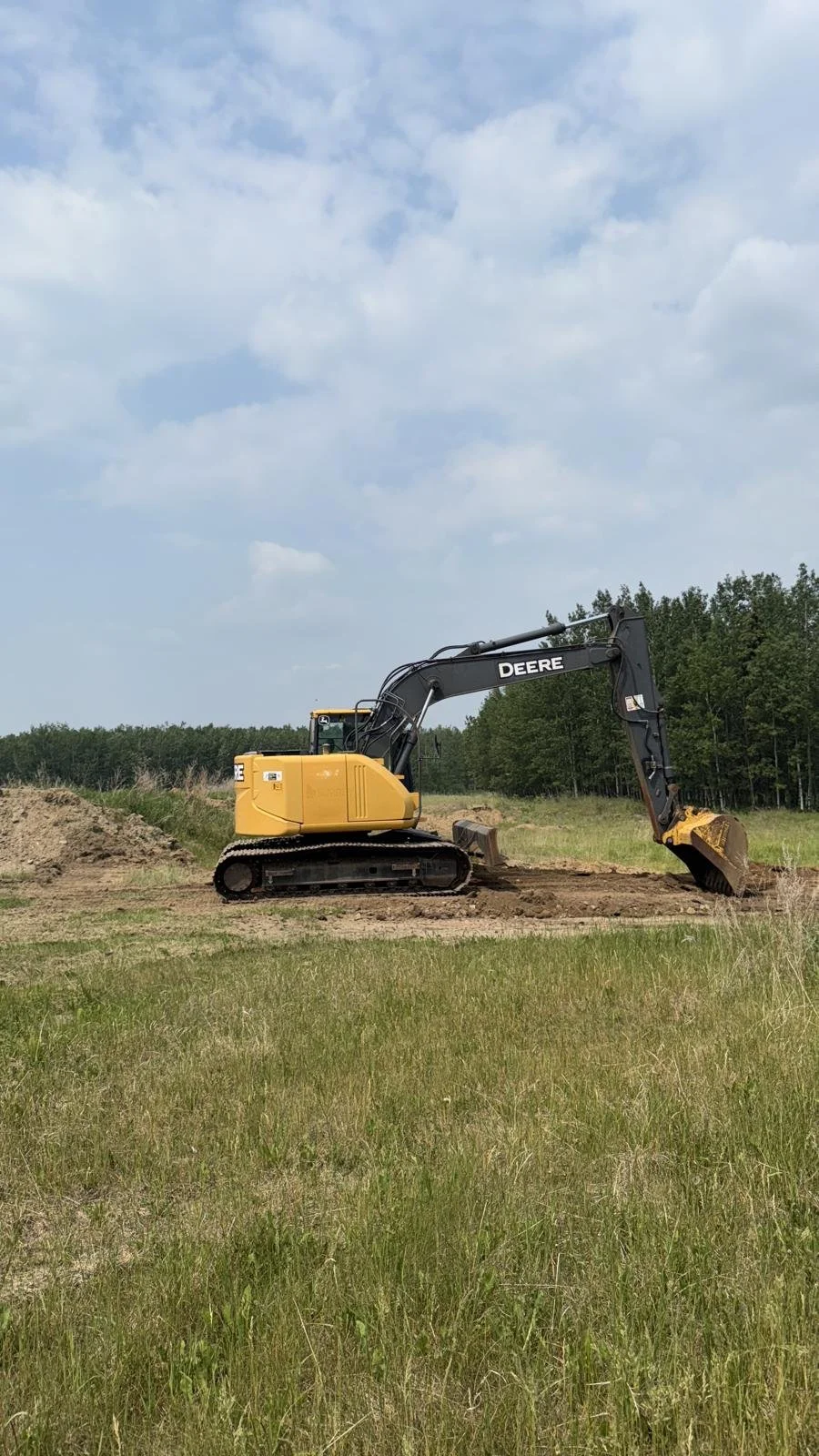 Yellow and black excavator working on land in a field with trees in the background under partly cloudy sky.