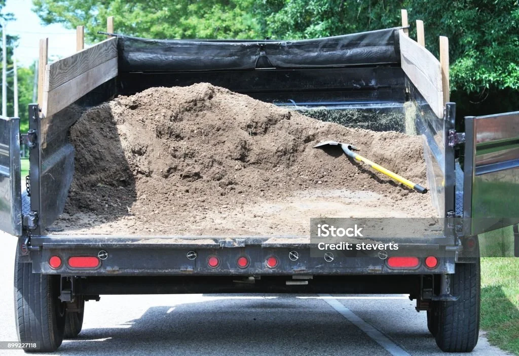 A black pickup truck with open bed carrying a large pile of dirt and a shovel inside.