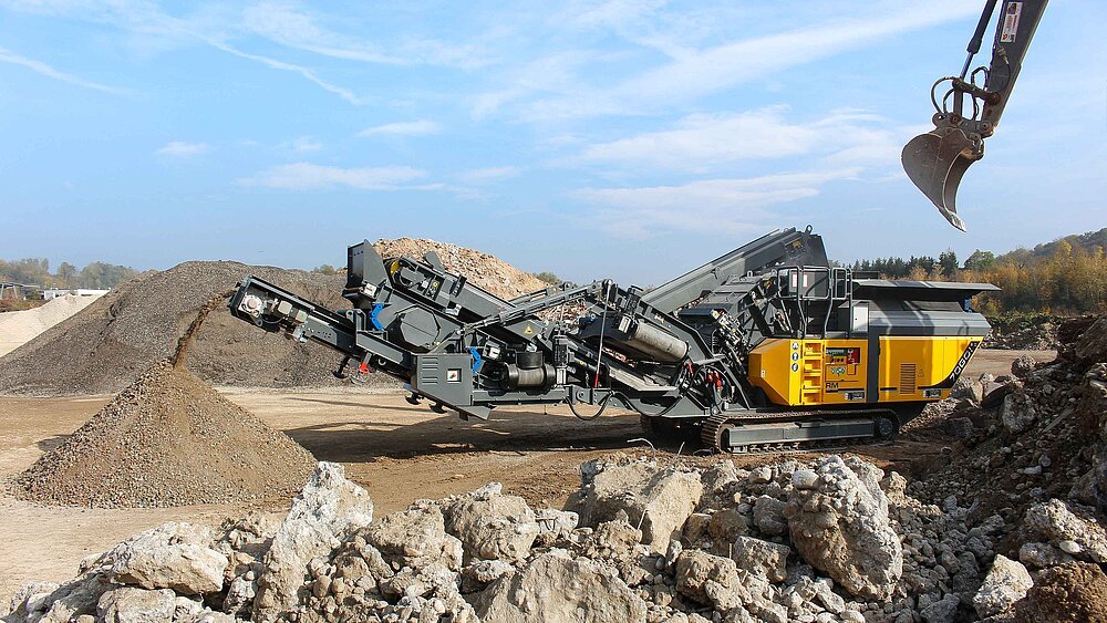 A large mobile rock crusher on a construction site, with rocks and gravel around it and a clear blue sky above.