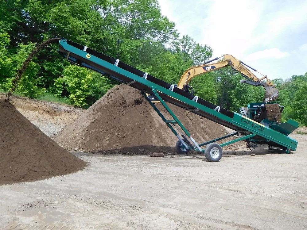 A large soil screener machine sorting dirt into piles on a construction site with trees in the background.
