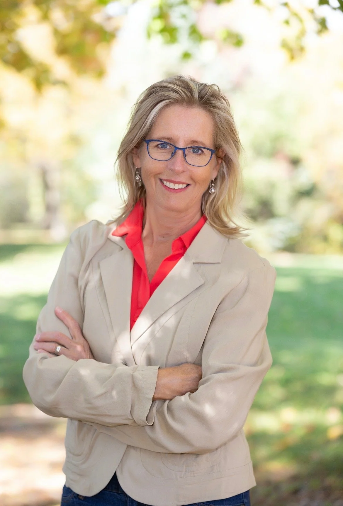 A woman with blonde hair, blue glasses, and a bright smile, wearing a beige blazer and red blouse, standing outdoors with trees and greenery in the background.