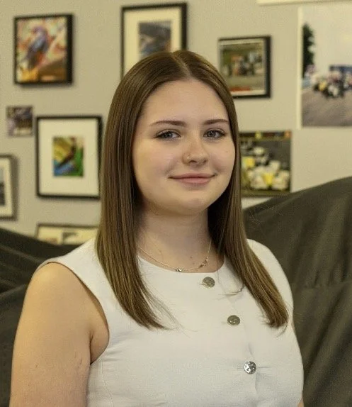 A young woman with straight, shoulder-length brown hair, wearing a sleeveless white top with silver buttons, and a small necklace, standing in an art gallery with framed pictures on the wall behind her.