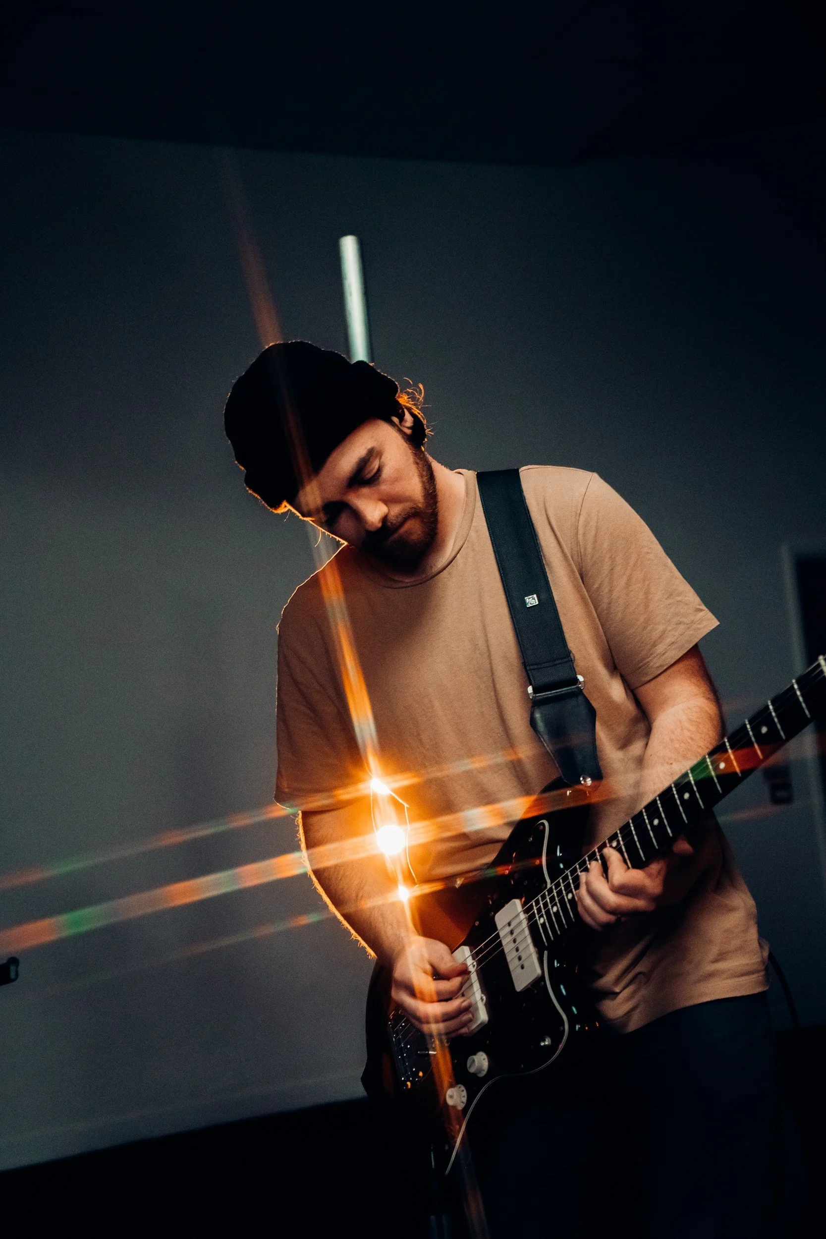 A man with dark hair, beard, and a black beanie playing an electric guitar in a dimly lit room with reflections of light.