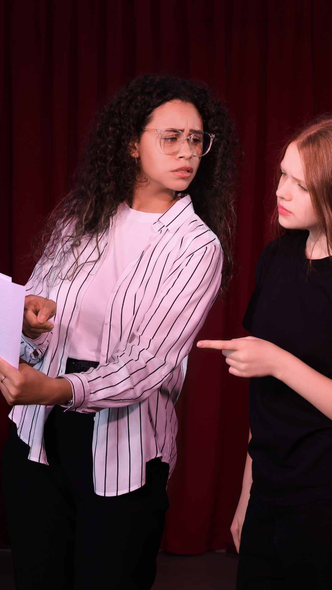 Two women having a heated argument, one with curly hair and glasses, holding a notebook, and the other with straight red hair pointing at her.