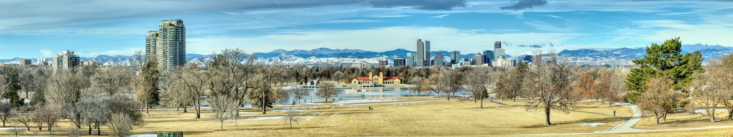 Panoramic view of Denver skyline with mountains in the background and a park with trees and a pond in the foreground.