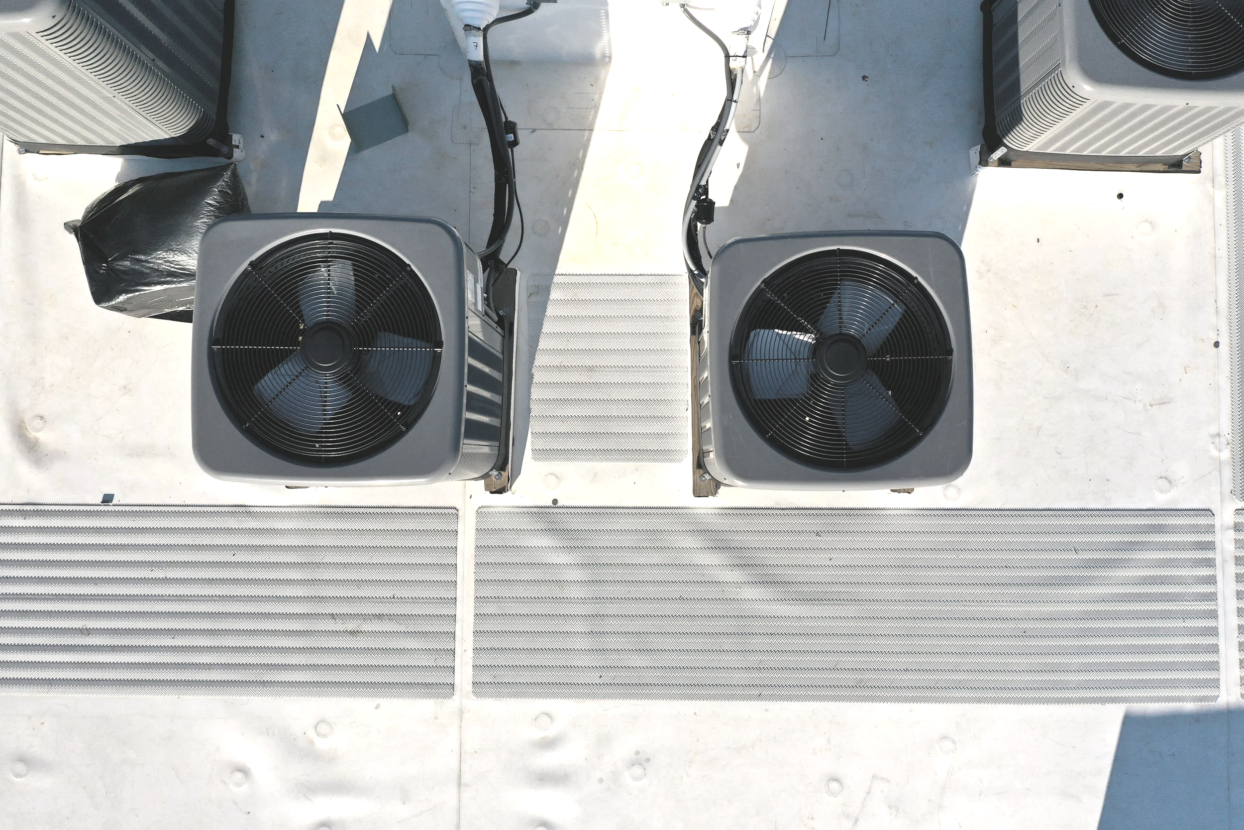 Two large outdoor air conditioning units with black fans on a rooftop with a white surface, electrical wiring, and a black plastic bag.