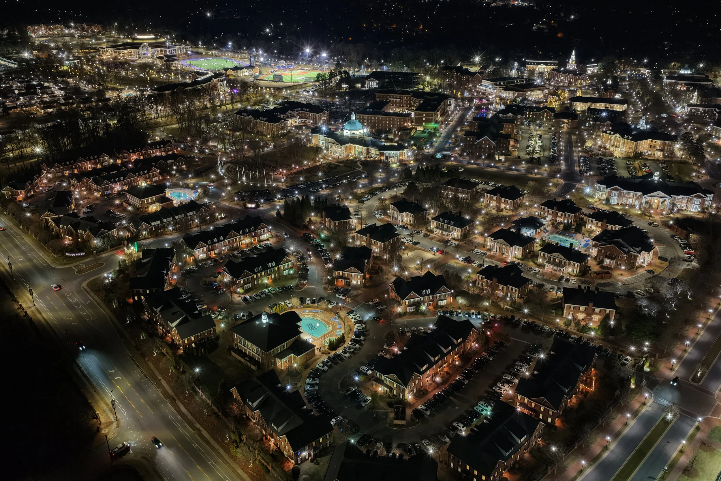 Night aerial view of a well-lit cityscape with residential complex, swimming pools, parking lots, and a sports stadium in the background.
