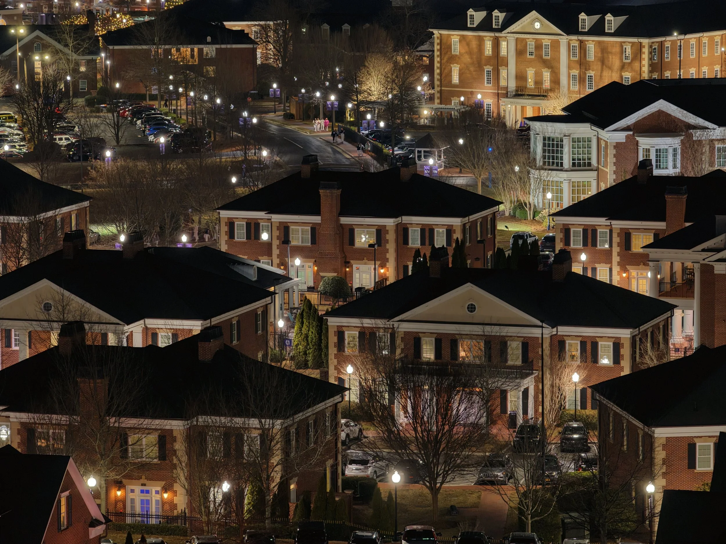 Night view of a residential neighborhood with multi-story brick houses, lit street lamps, leafless trees, and parked cars.