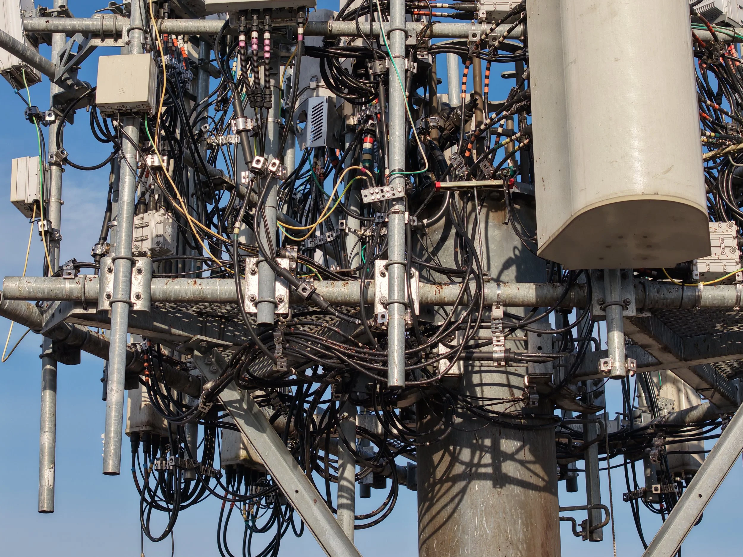 Close-up of a utility pole with numerous tangled black wires, electrical boxes, and metal supports against a blue sky.