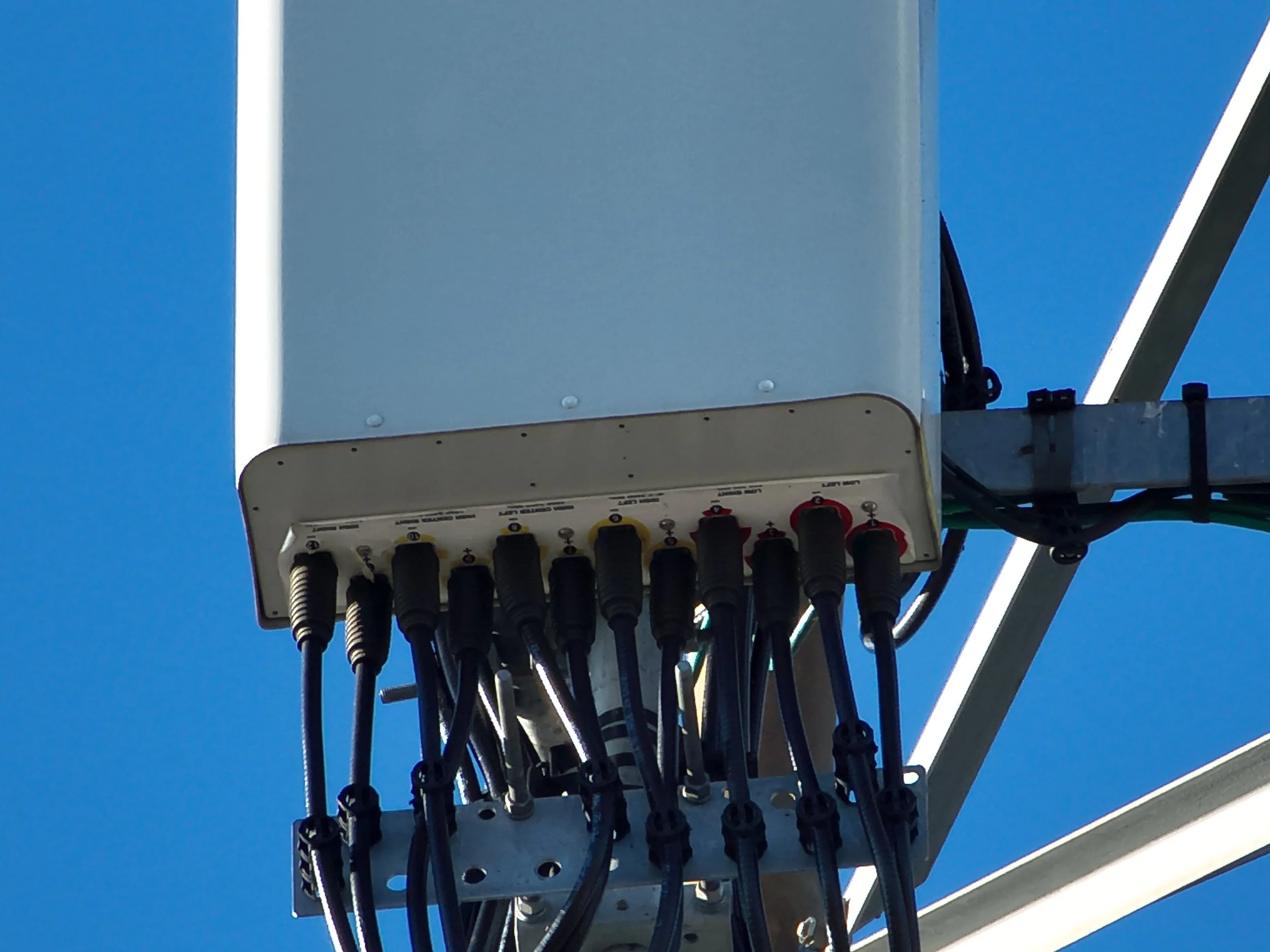 Close-up of a telecommunications antenna tower with various cables connected at the base of the antenna structure against a clear blue sky.