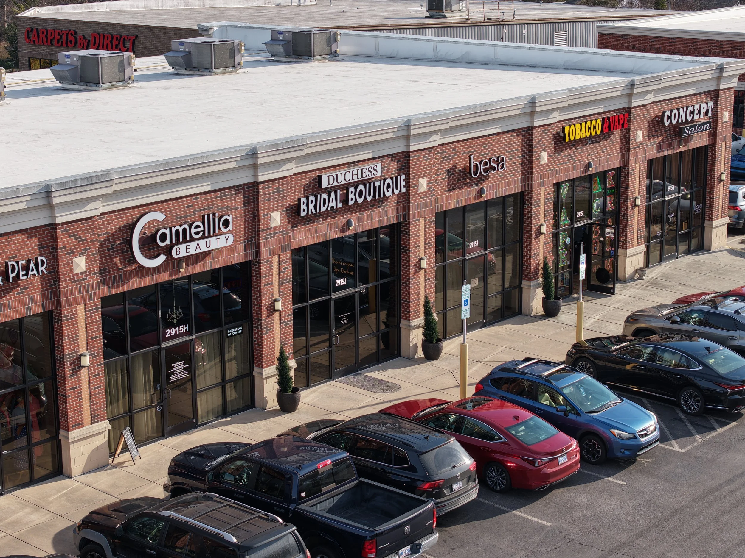 A strip mall with various retail stores, including a beauty salon, bridal boutique, and vape shop, with cars parked in front and potted plants outside the entrances.