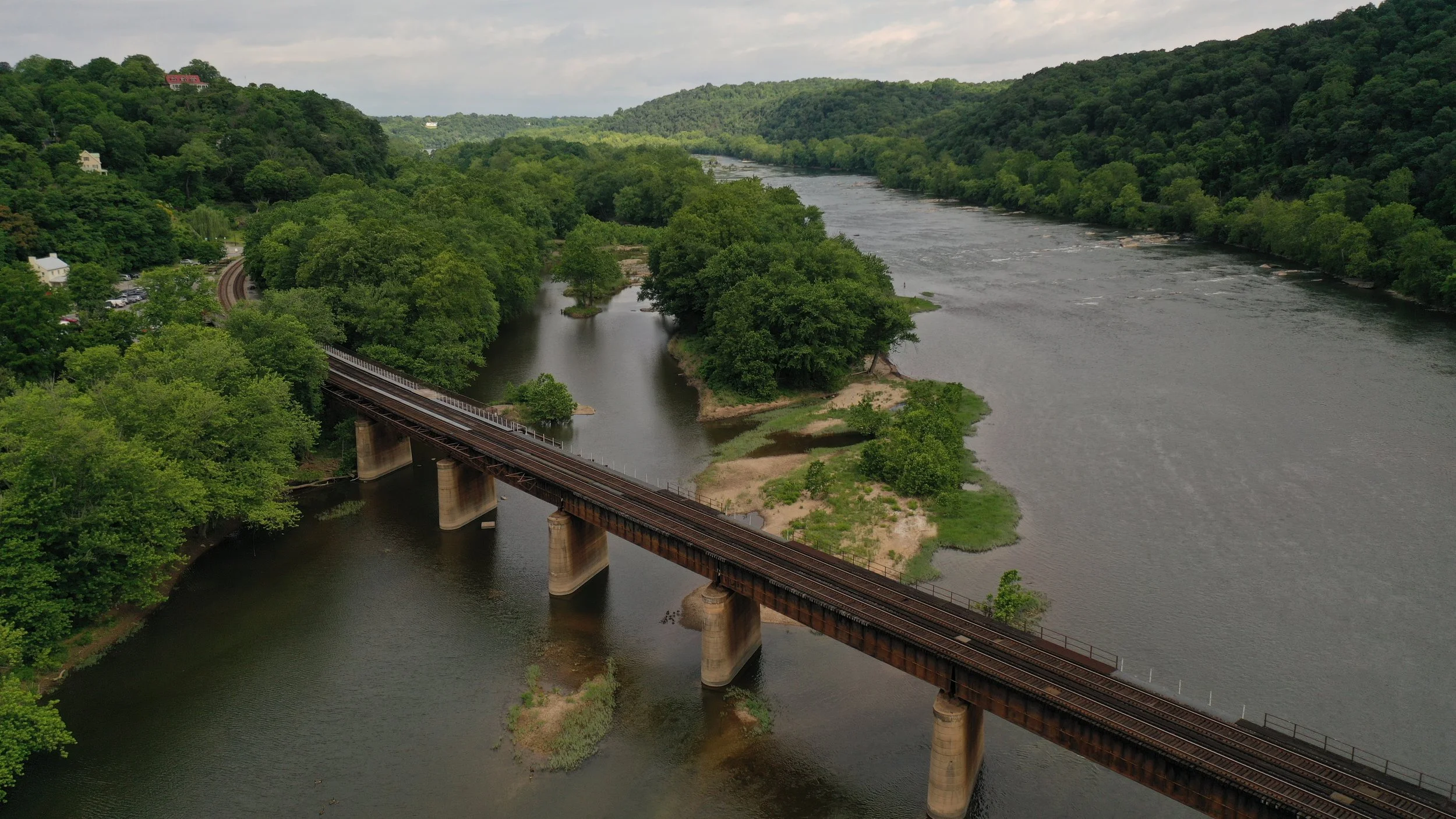 Aerial view of a railroad bridge crossing a river, with lush green trees and hills on either side.
