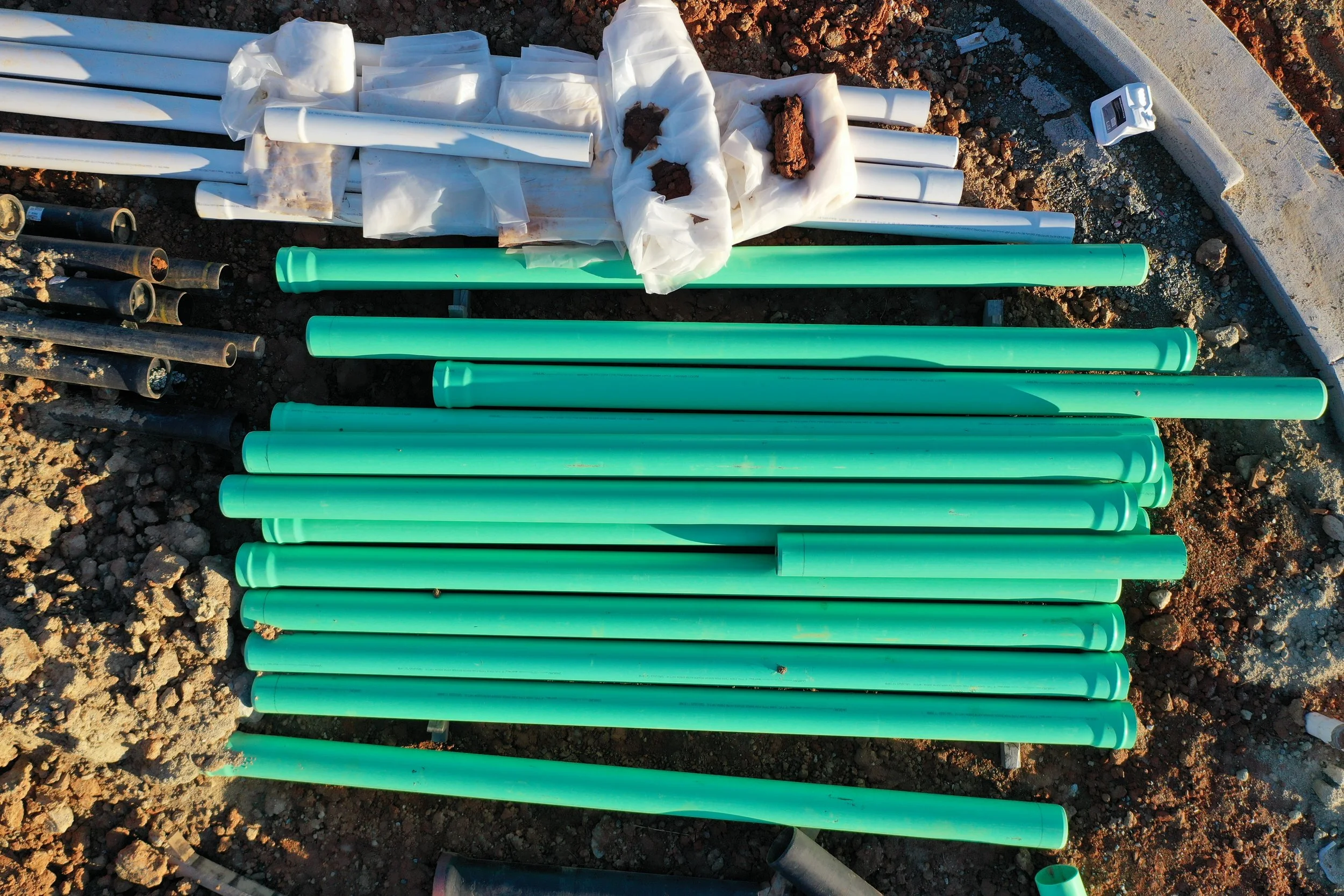 Stacked green PVC pipes and white packaging on dirt ground at a construction site.