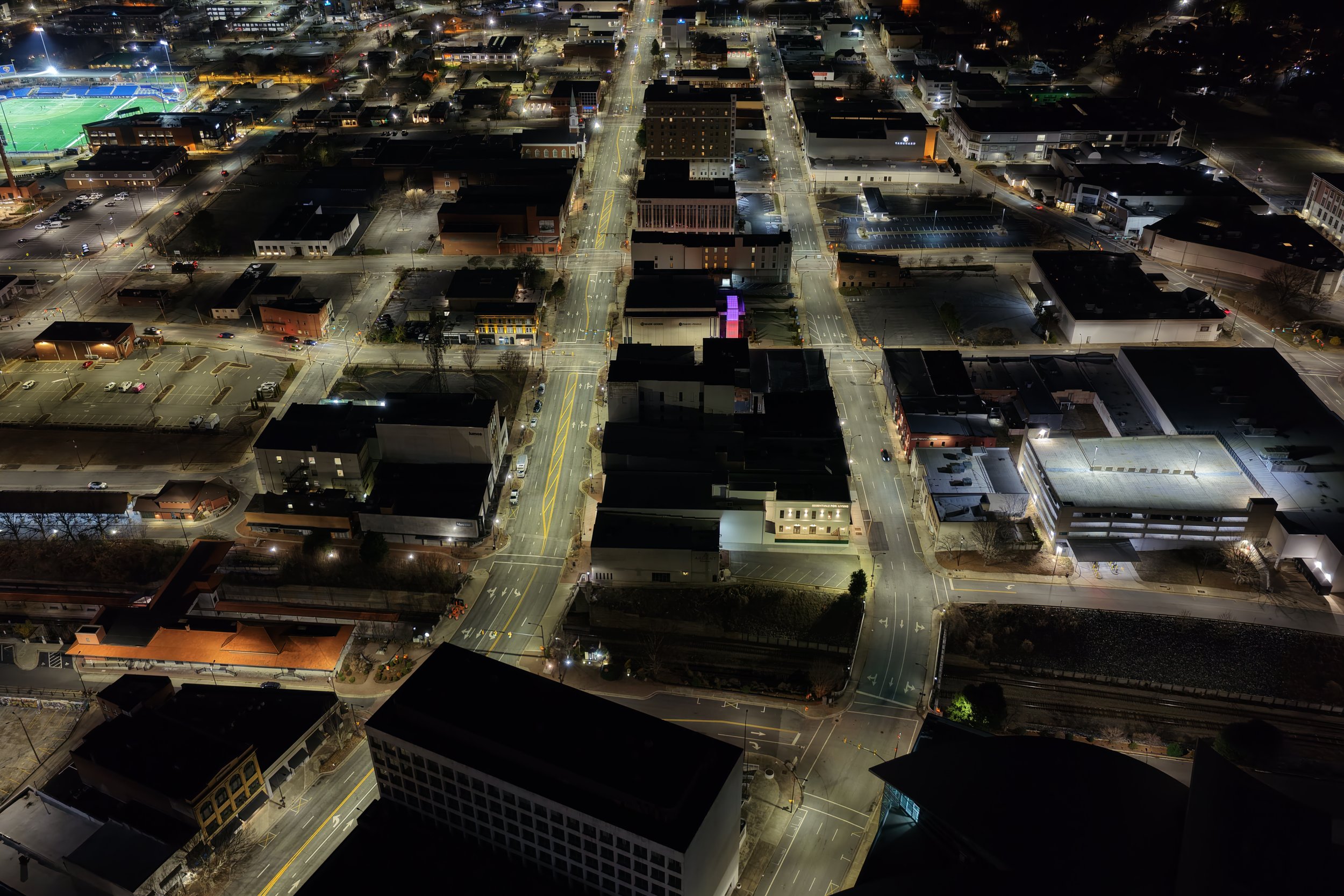 An aerial view of a city at night, showing illuminated streets, buildings, parking lots, and a sports field with green grass in the upper left corner.