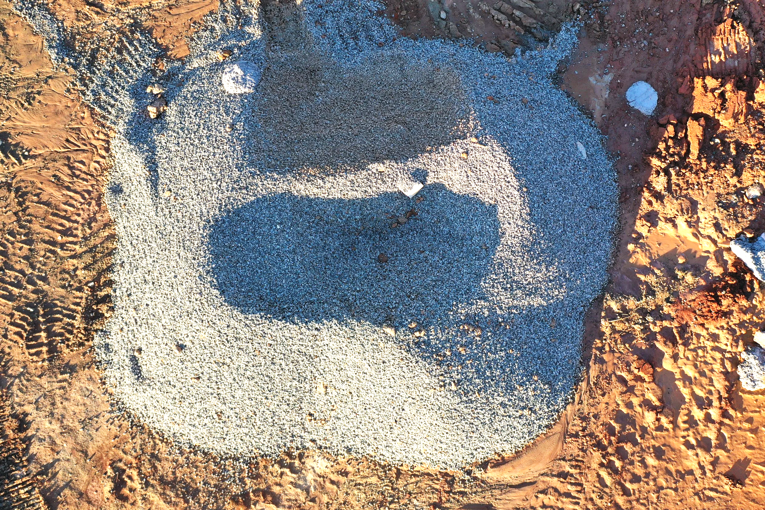 A bird's-eye view of a large heart-shaped sand sculpture on a sandy beach, with tire tracks nearby and some scattered rocks.