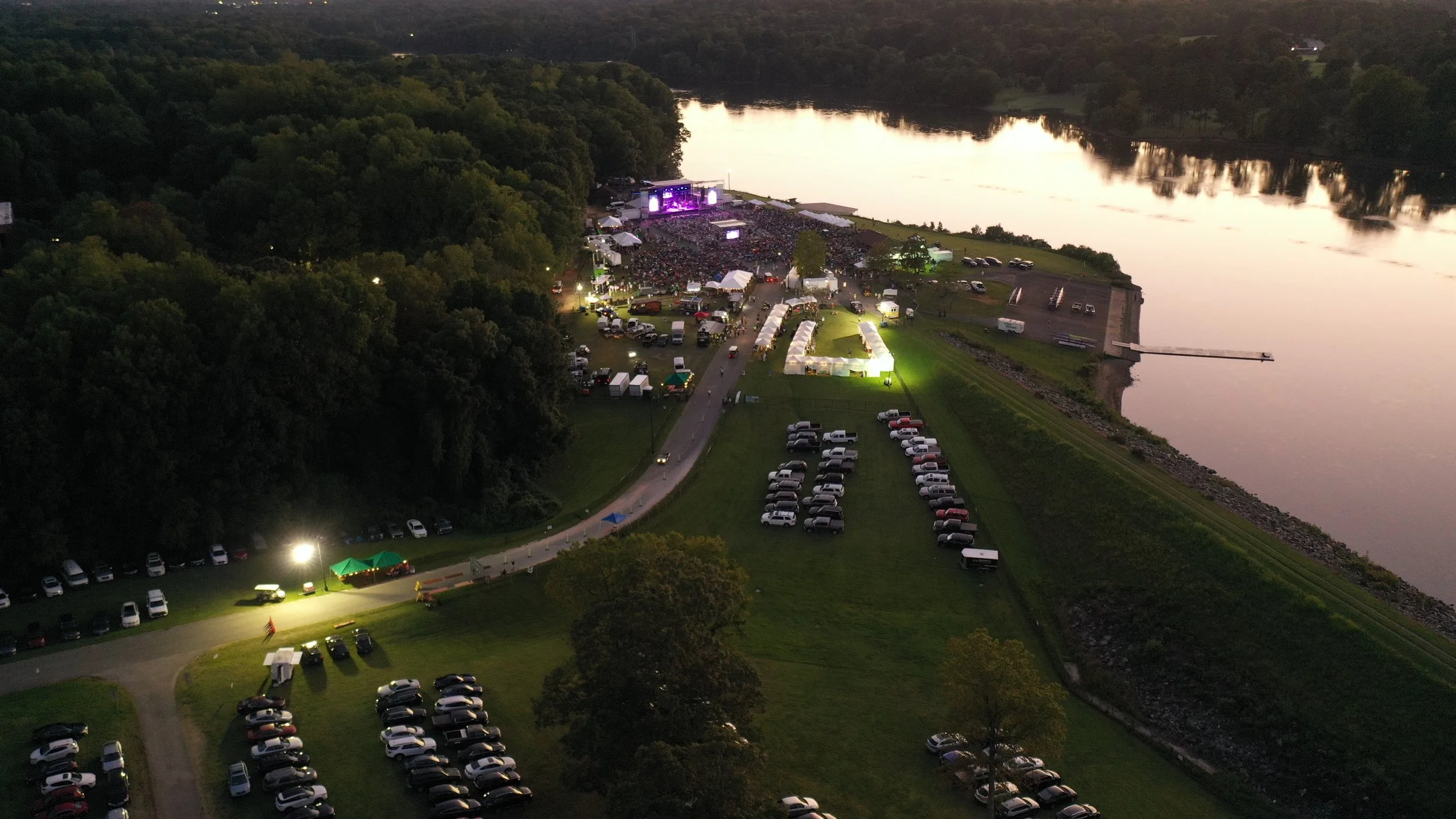 An aerial view of a large outdoor concert by a river at dusk, with a stage, crowd, tents, and parked cars nearby.