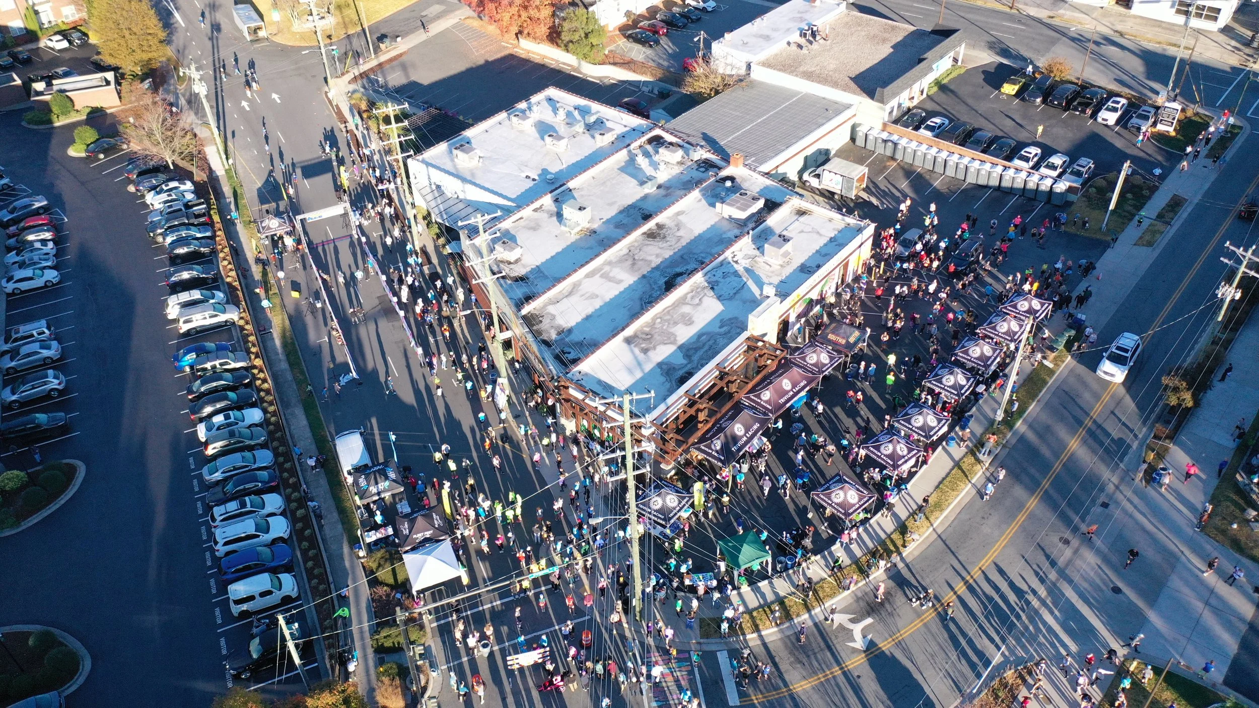 Aerial view of an outdoor event with a large crowd gathered around a building with a white roof, tents, and booths, along with adjacent parking lots and streets.