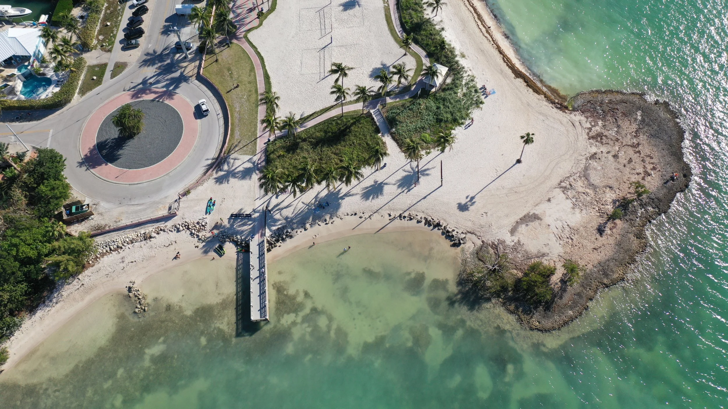 Aerial view of a small beach with a pier, surrounded by rocky shoreline, park area with trees, and nearby parking lot.