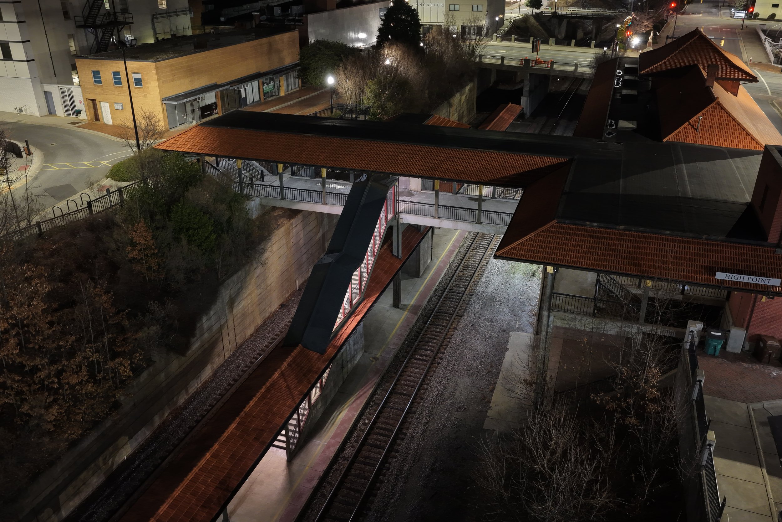 Night view of a train station platform with covered walkway and train tracks, overhead pedestrian bridge, surrounding buildings, and trees.
