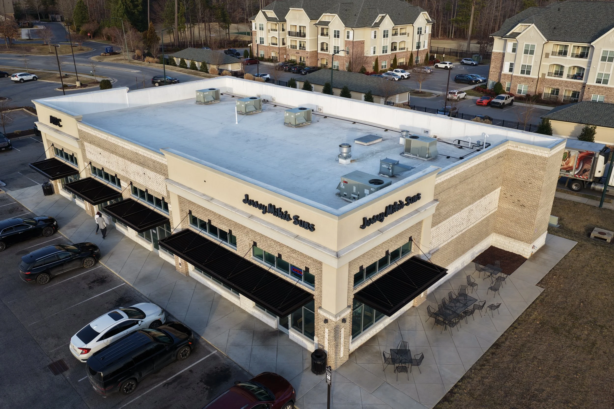 Aerial view of Jersey Mike's Subs restaurant in a strip mall, showing parking lot, outdoor seating, and surrounding residential area.