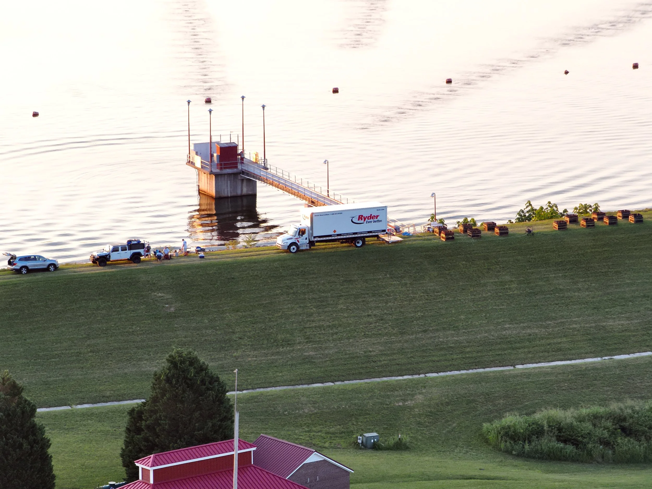 A lake with a dock, several vehicles on the grassy shore, and a row of seating areas. The scene is viewed from above during the daytime.