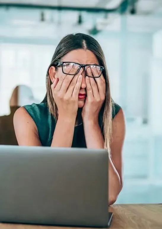 A woman with glasses sitting in front of a laptop, holding her face in her hands, appearing stressed or overwhelmed.