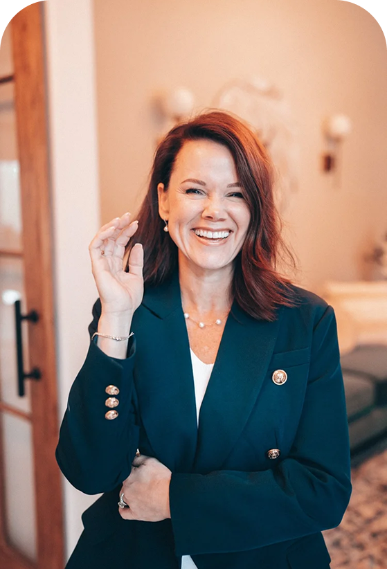 A woman with red hair laughing and smiling, wearing a navy blazer with gold buttons, in a warmly lit indoor space.