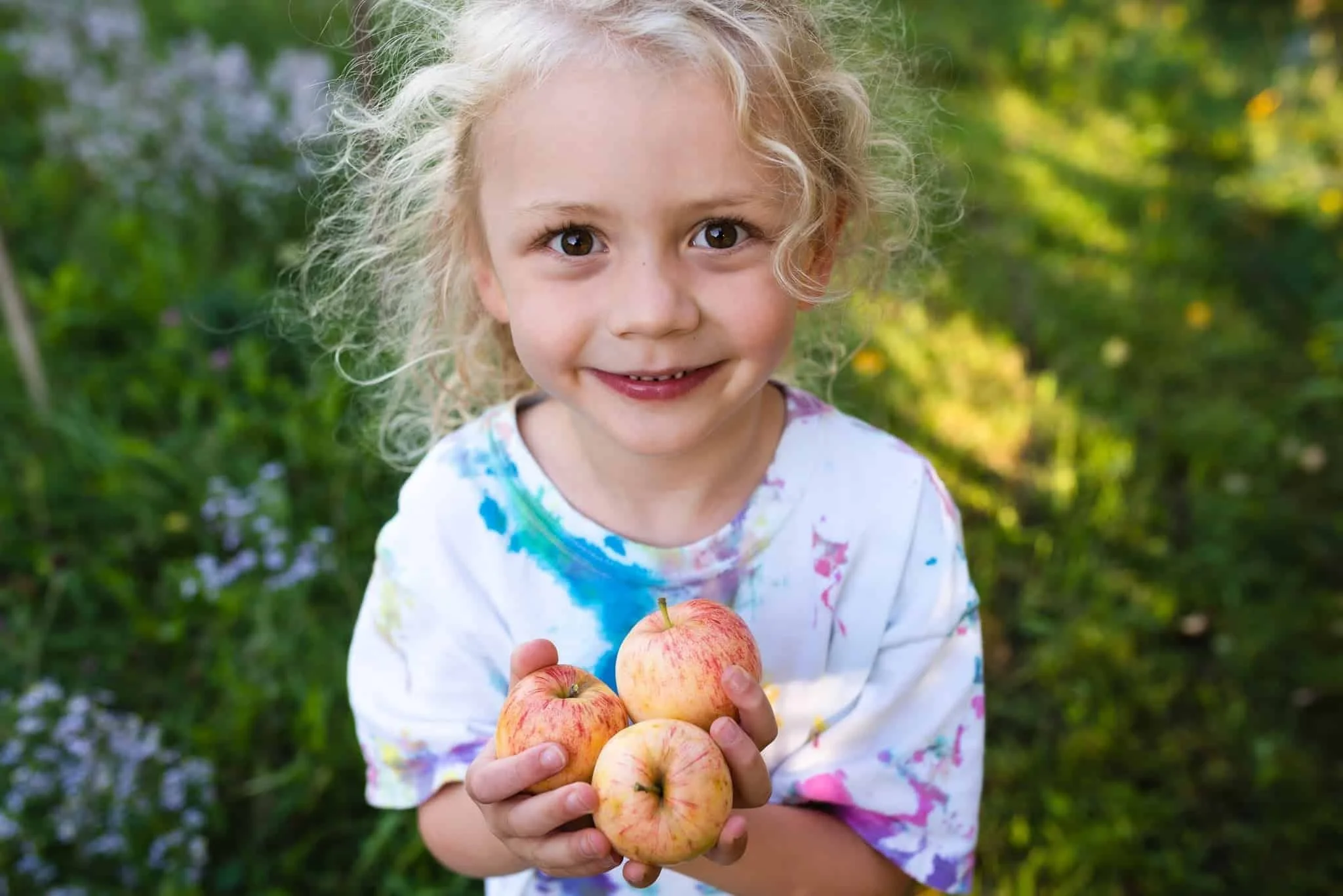 Boy smiling with blond hair and three apples in his hands from food forest garden.