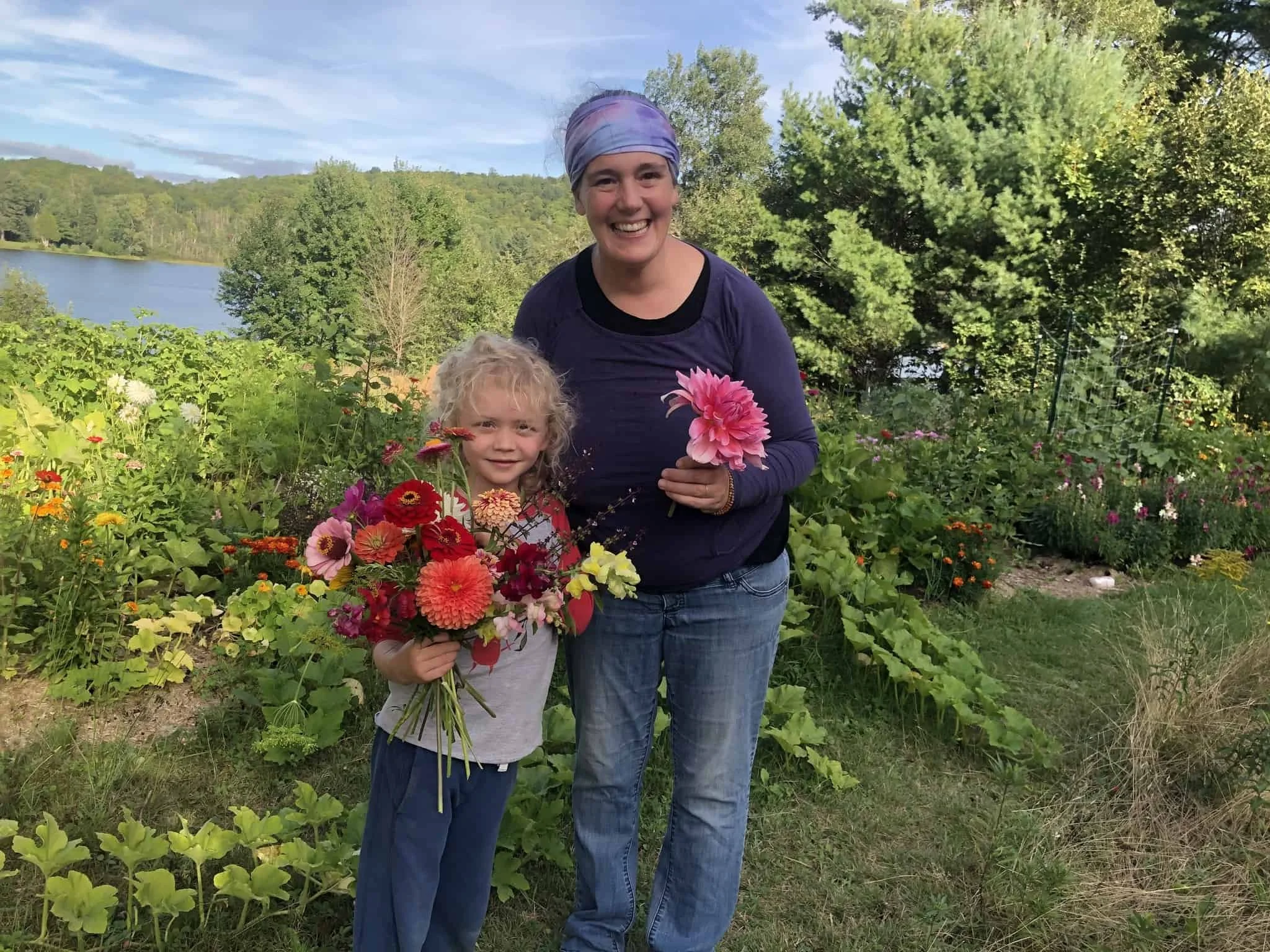 Mom and son standing in food forest holding huge colourful flower bouquets made of dahlias, zinnias, snap-dragons and basil.